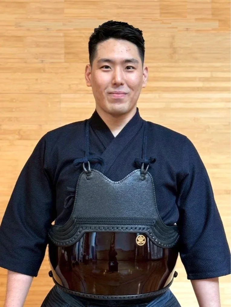 Young man in black martial arts uniform wearing a chest protector against a wooden wall.