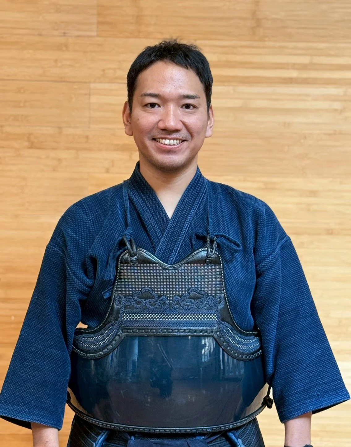 Man in traditional martial arts uniform wearing protective gear, standing indoors in front of a wooden wall.
