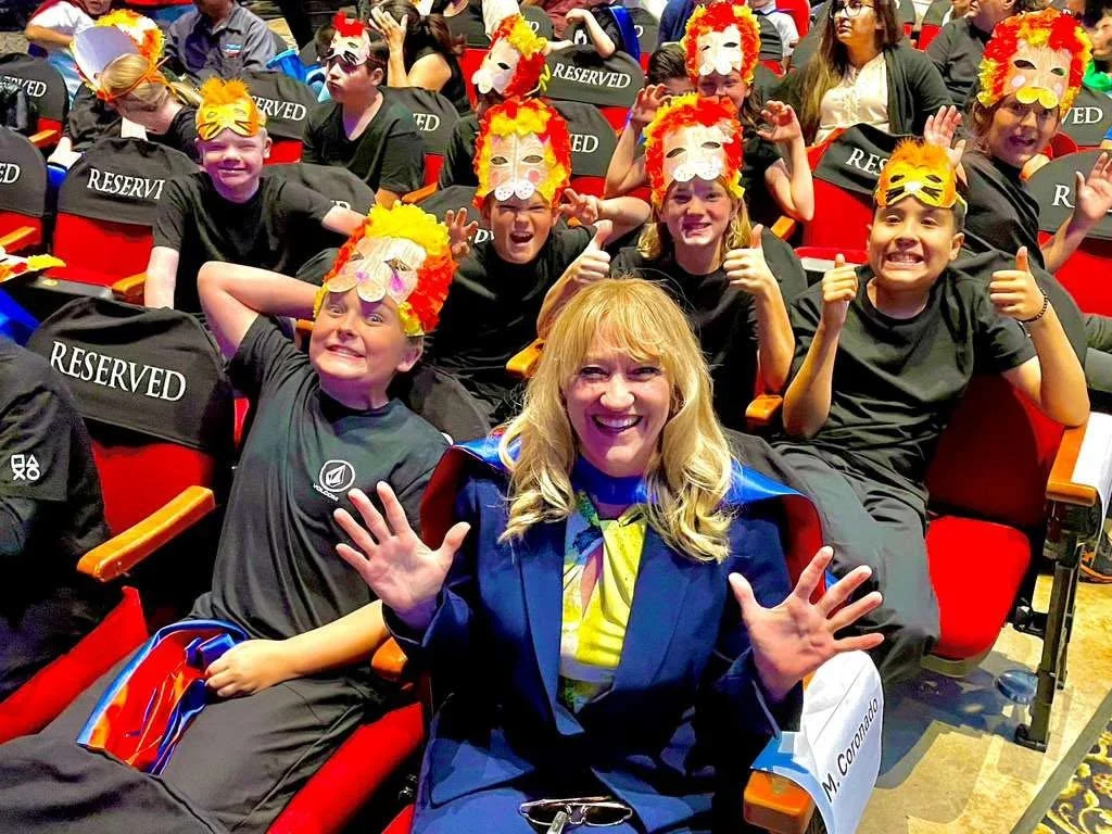 Cali sitting in the audience holding her hands up and smiling while sitting with middle school students.