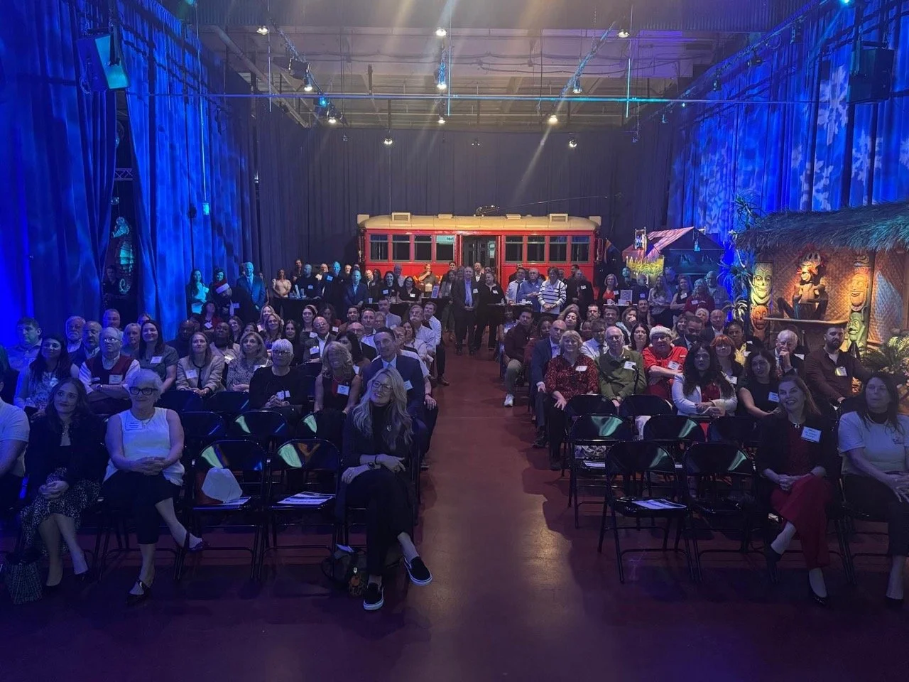 Crowd of adults sitting in a room in front of a red train cart at the Meet and Greet Cali event at Garner Holt Productions.