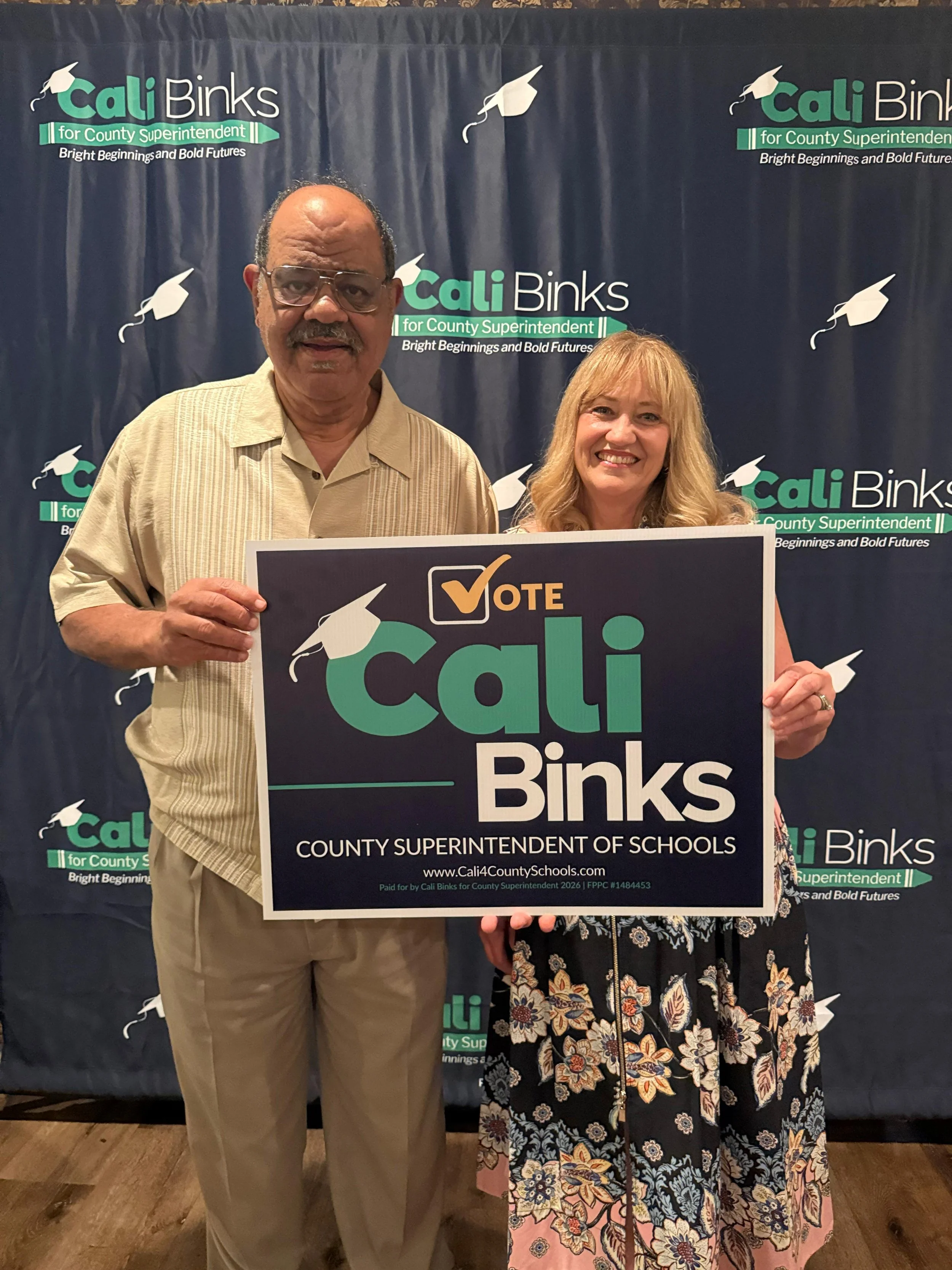 Cali Binks next to a guest holding a yard sign in front of a step and repeat background.