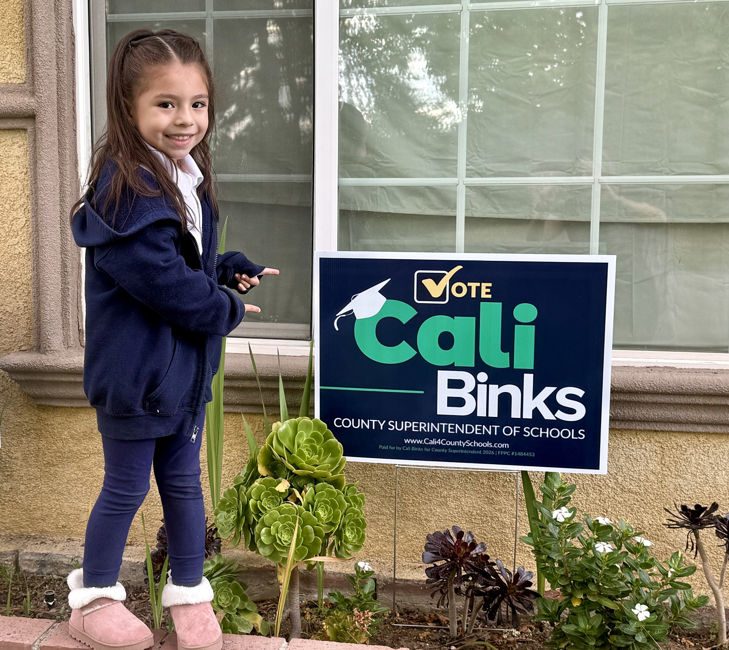 5 year old child pointing to a campaign yard sign saying, "Vote Cali Binks, County Superintendent of Schools, www.cali4countyschools.com"
