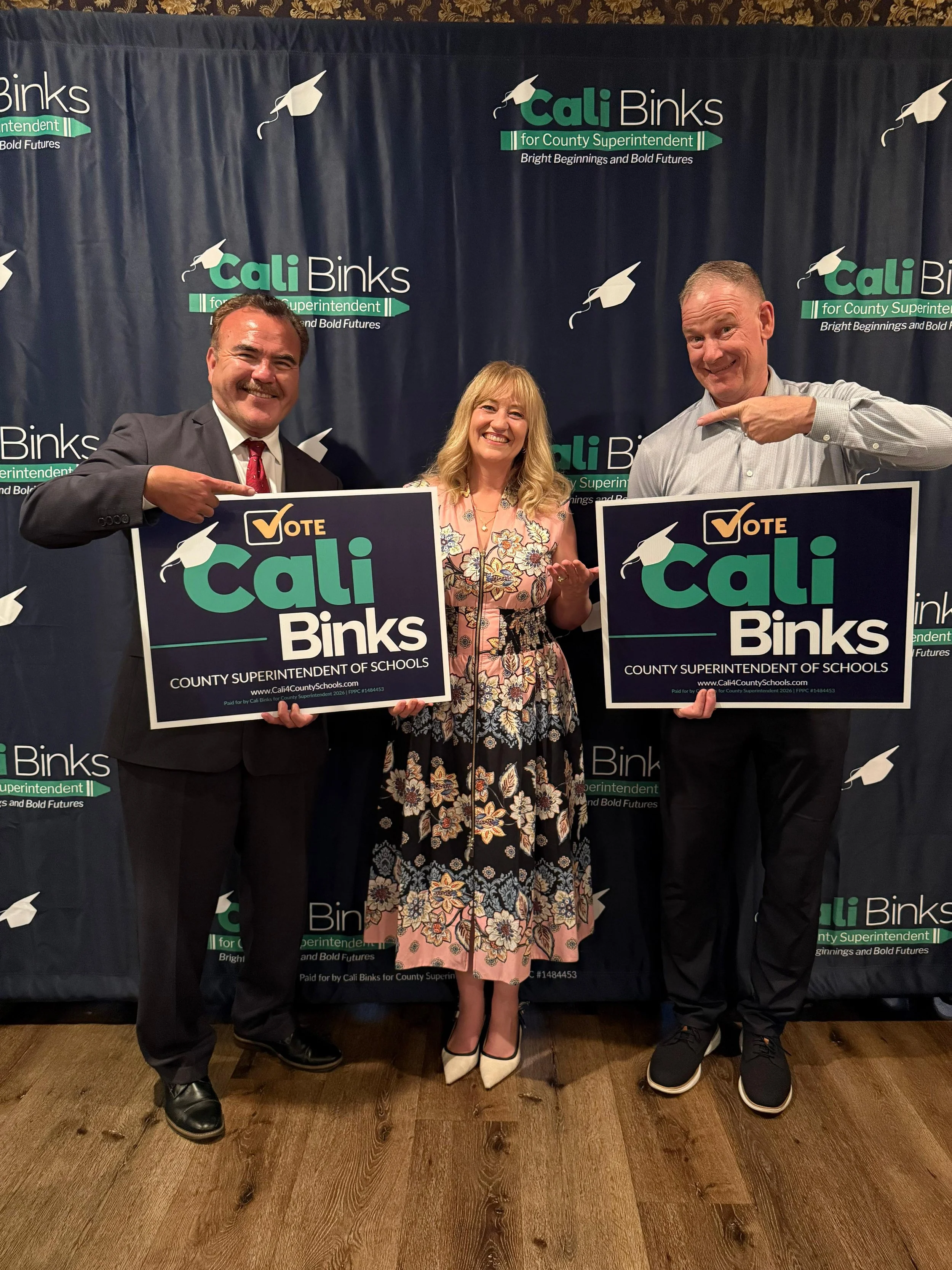 Cali Binks next to Ted Alejandre and a guest holding yard signs in front of a step and repeat background.
