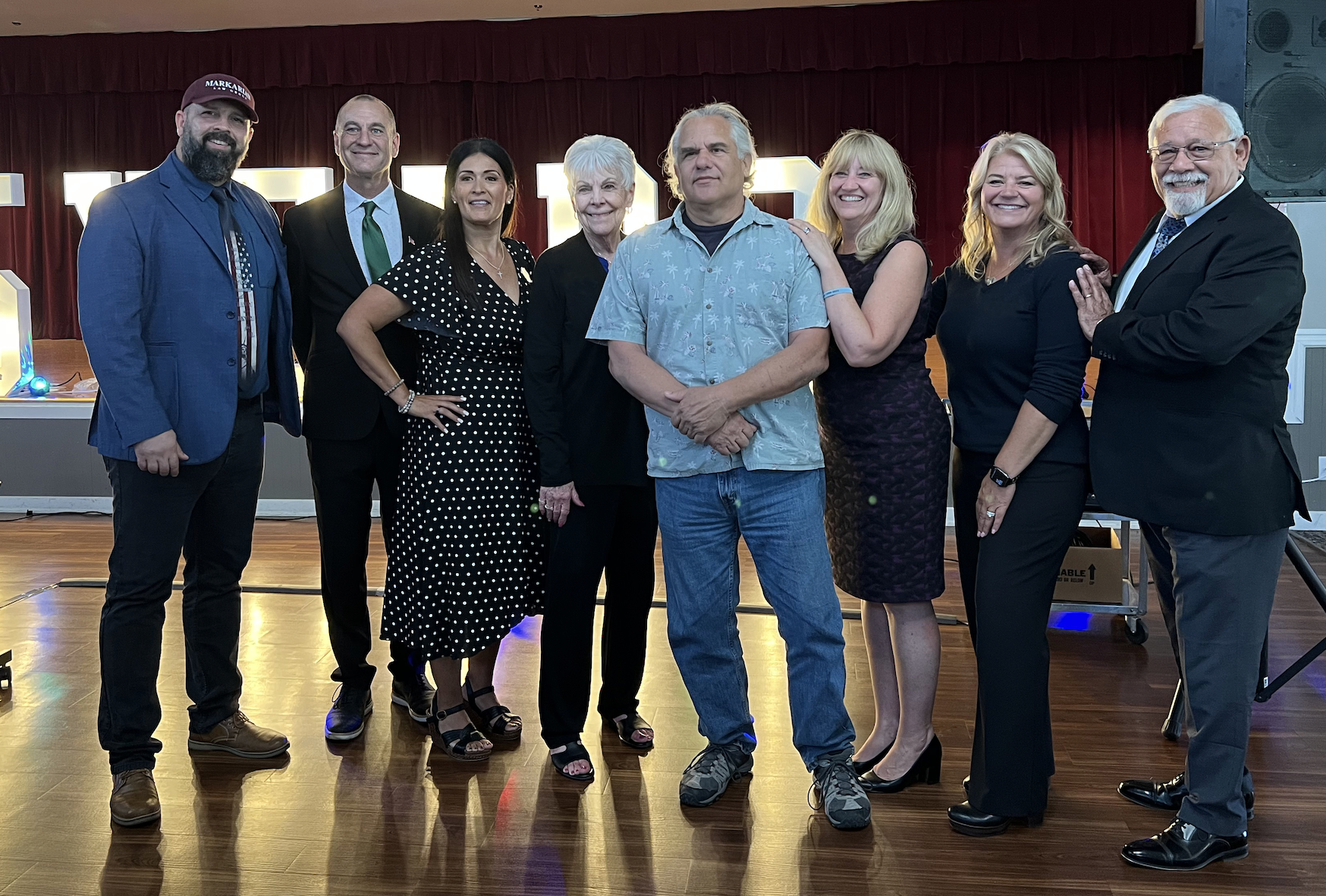 Seven adults standing in front of a stage surrounding Cali at a local event.