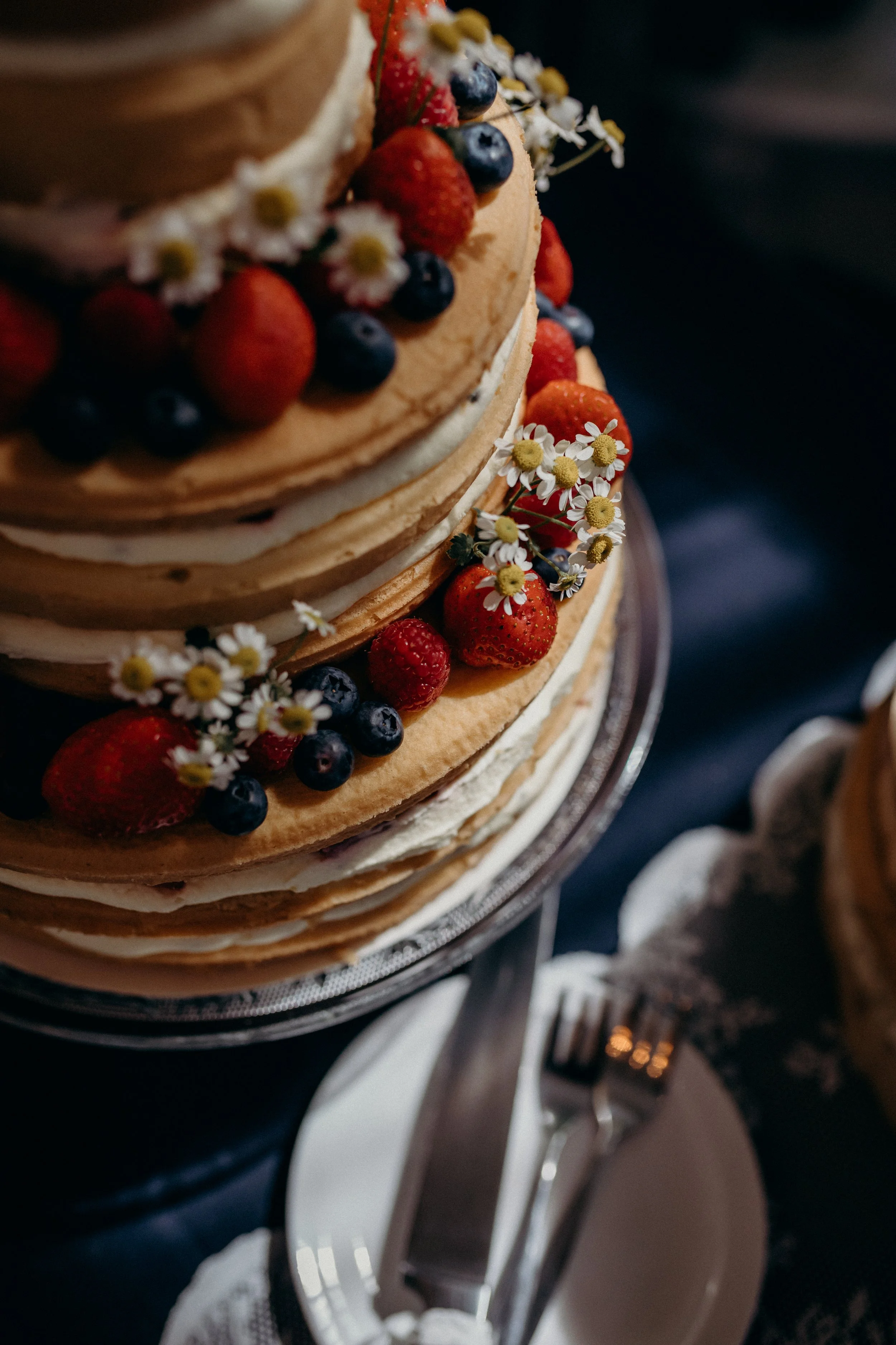 A multi-layered naked cake decorated with fresh strawberries, blueberries, raspberries, and small daisies, on a silver cake stand.