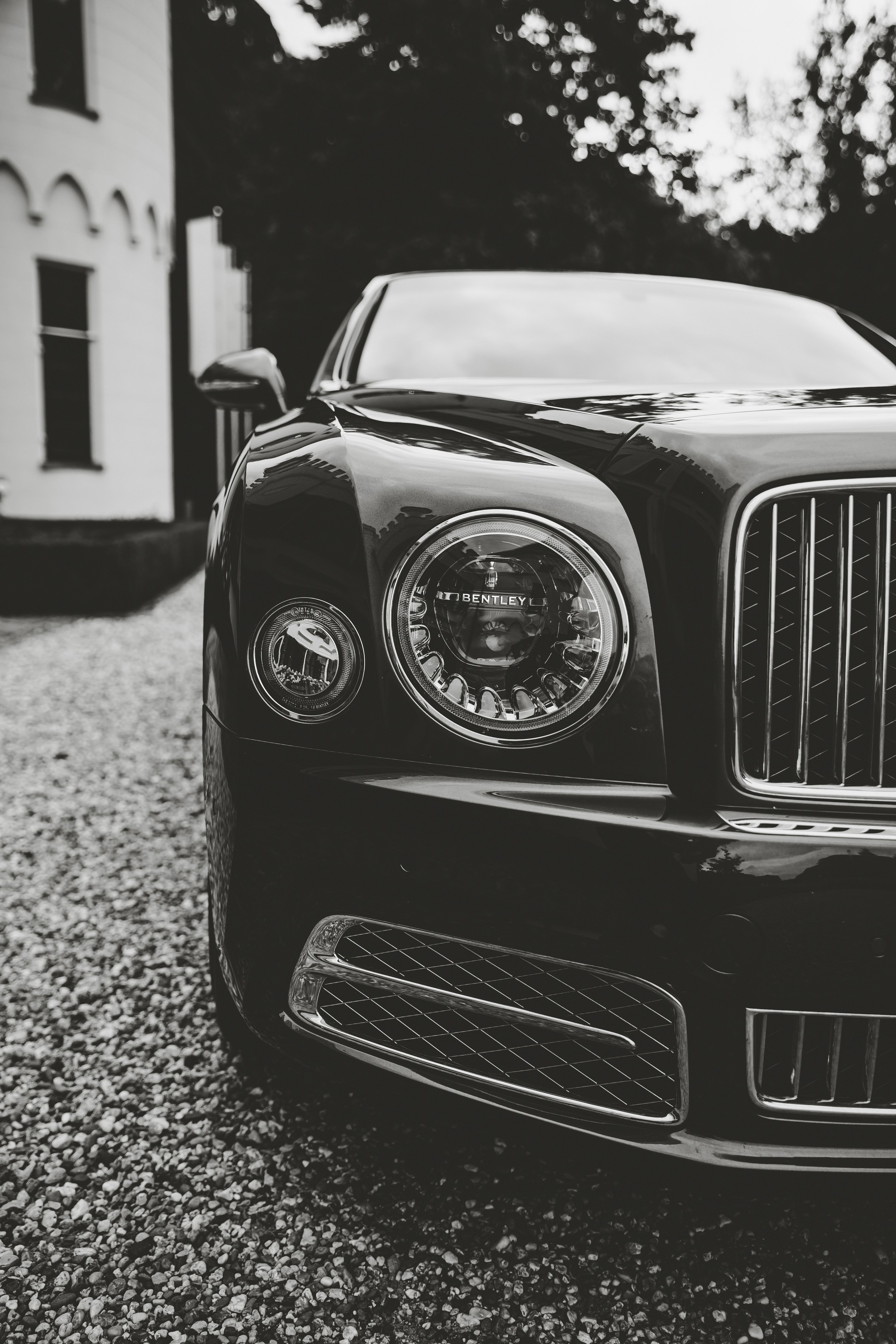 Close-up of front left side of a black Bentley car parked on gravel, showing the headlight, grille, and part of the bumper in black and white.