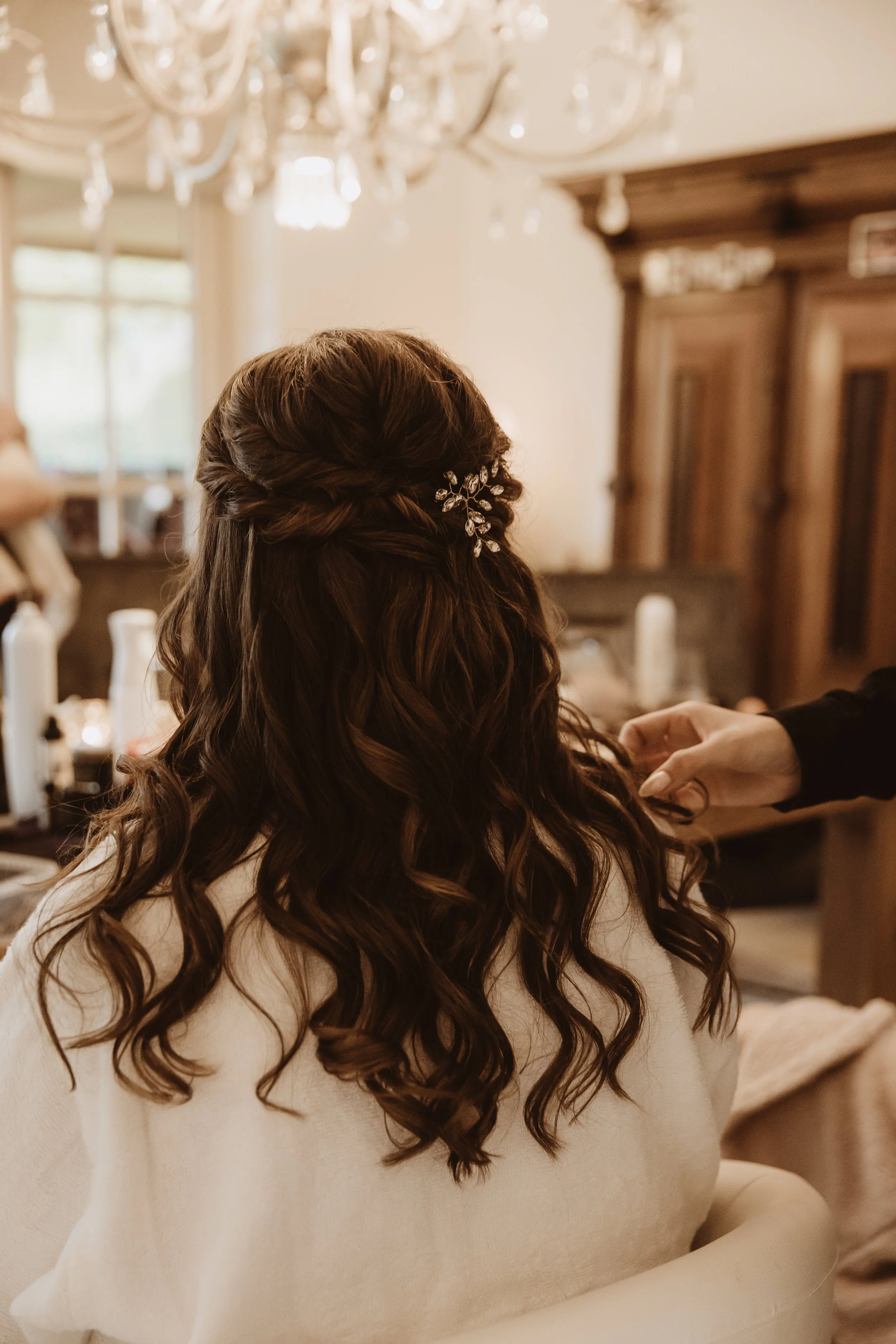 A woman with long, wavy brown hair sitting in a salon chair, having her hair styled with a decorative hairpin in an elegant, warmly lit room.
