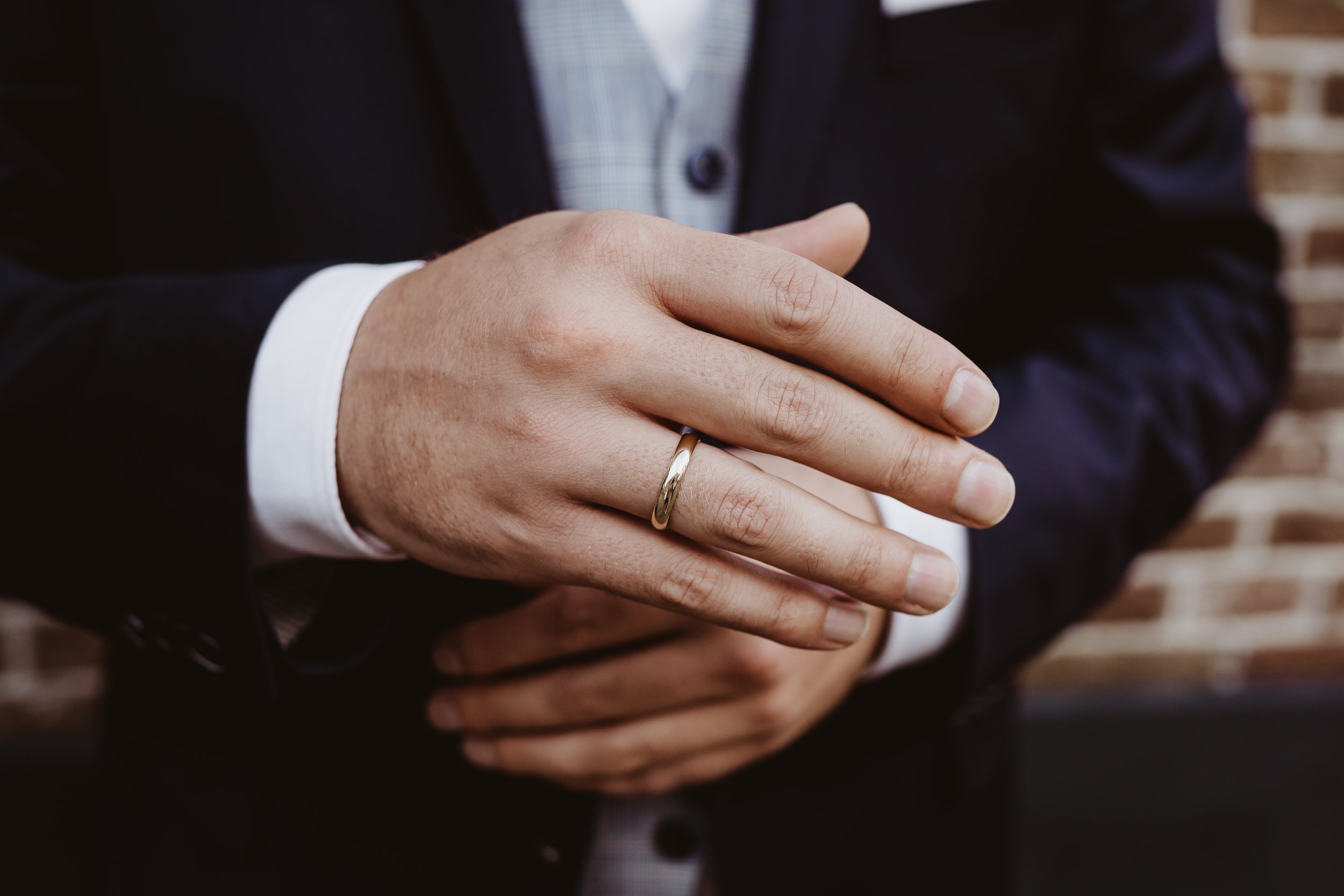 Close-up of a man wearing a wedding band on his ring finger, dressed in a dark suit, with hands crossed over his chest.