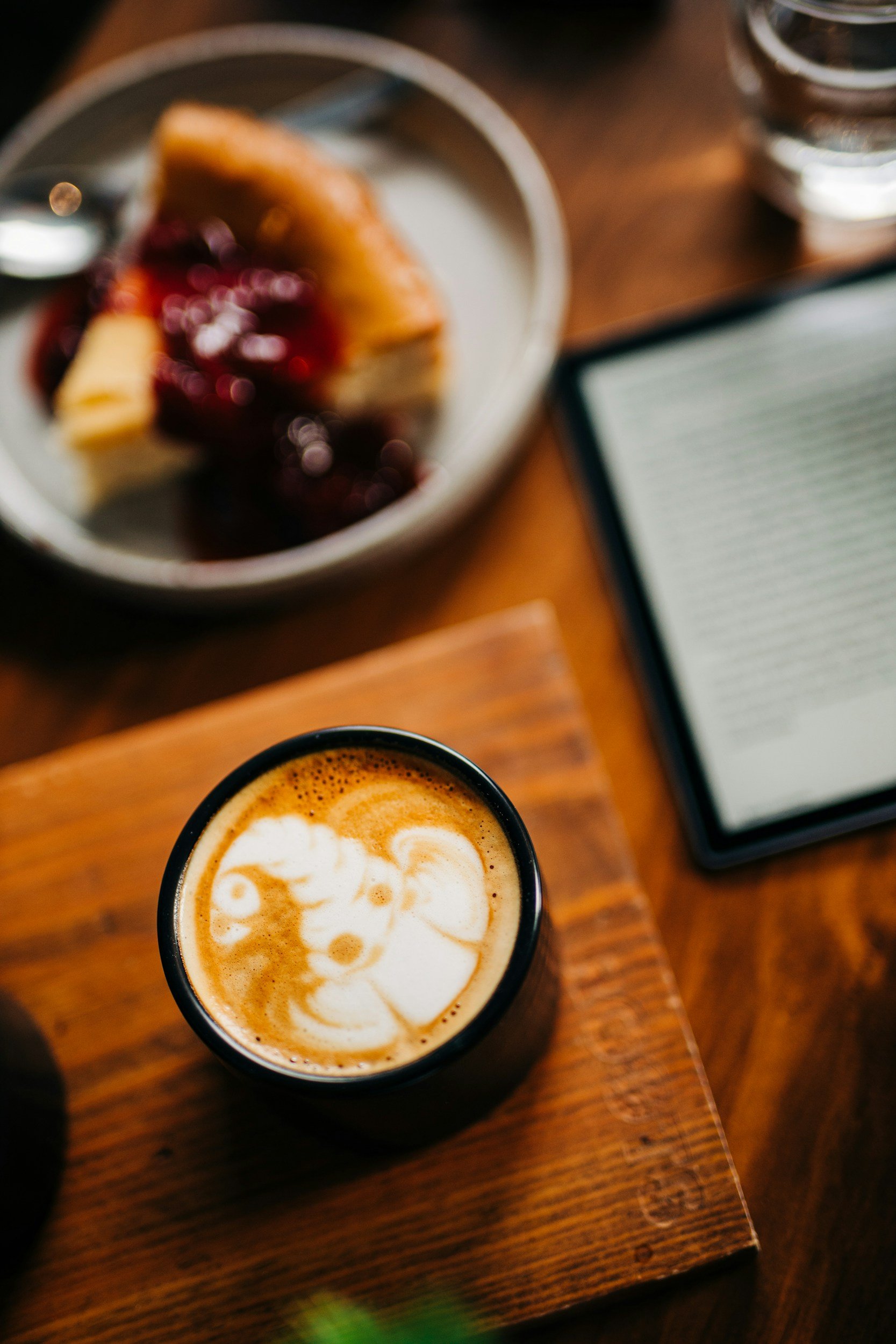 Close-up of a cup of coffee with latte art, placed on a wooden surface, with a blurry plate of cheesecake and a glass of water in the background.