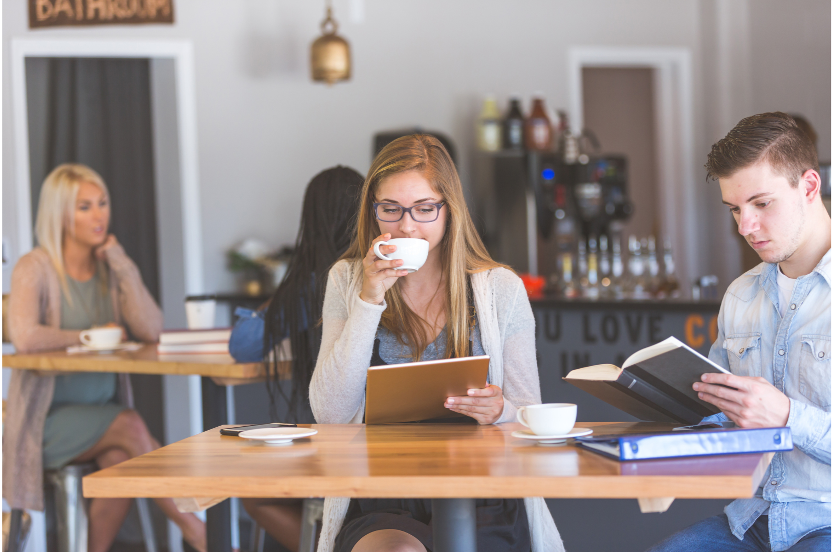A group of young people sitting at a wooden table in a coffee shop, engaging in different activities such as drinking coffee, reading, and talking.