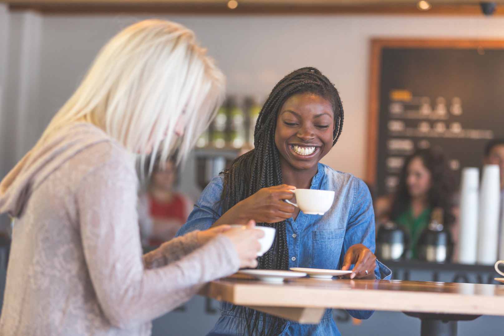 Two women enjoying coffee and conversation at a cafe, smiling and holding cups, with a menu board in the background.