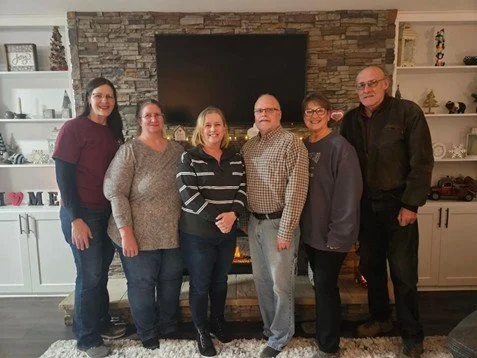 Six people standing in front of a fireplace with a flat-screen TV mounted above it, inside a room with white bookshelves and kitchen cabinets.