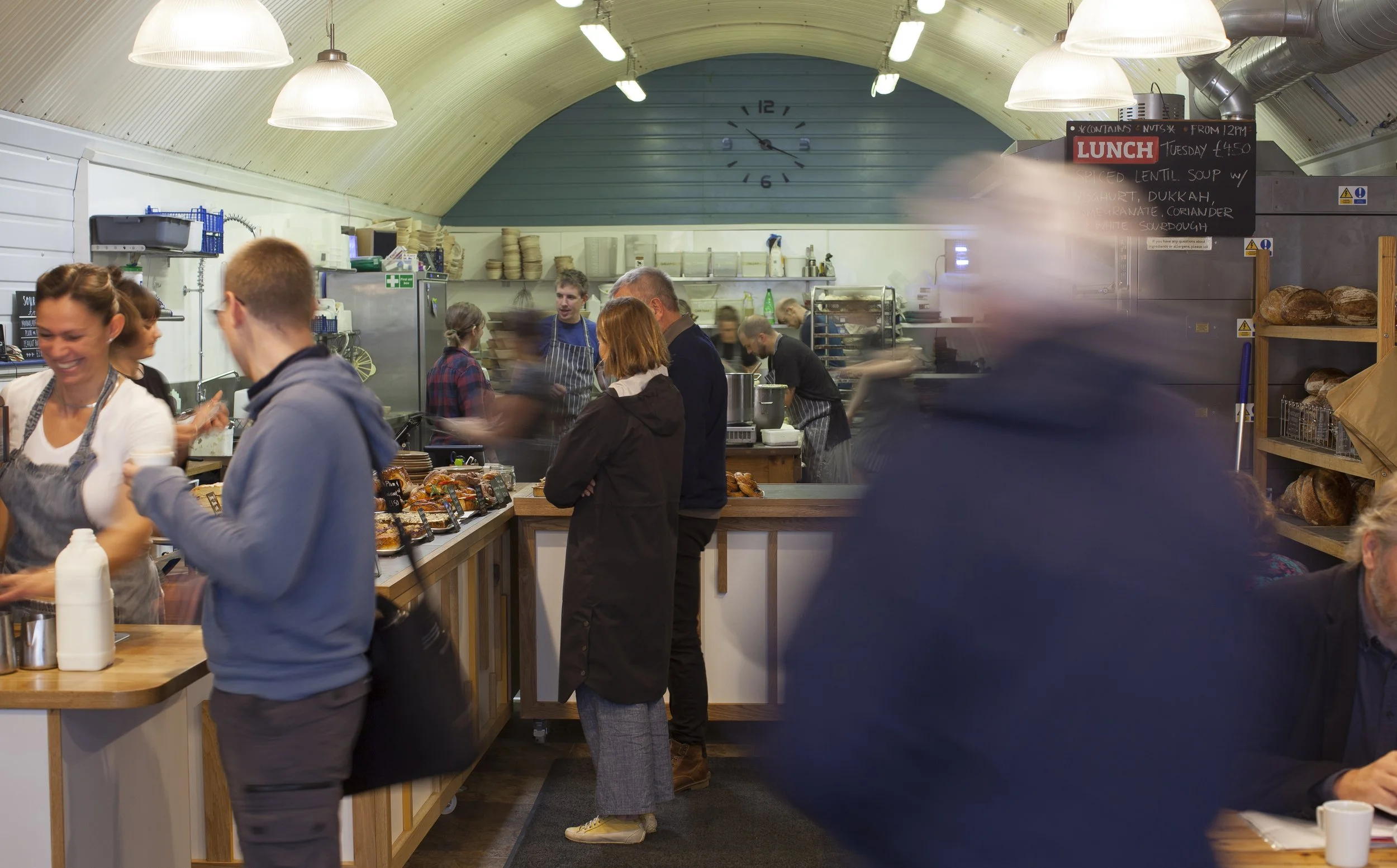 Inside Hart's Bakery in Bristol. Customers are waiting for their orders, a buffet of baked goods and a lady on the bottom left serving a customer, laughing with a pint of milk. Published as part of In Good Company series by The Lonely Hearts Club LHC