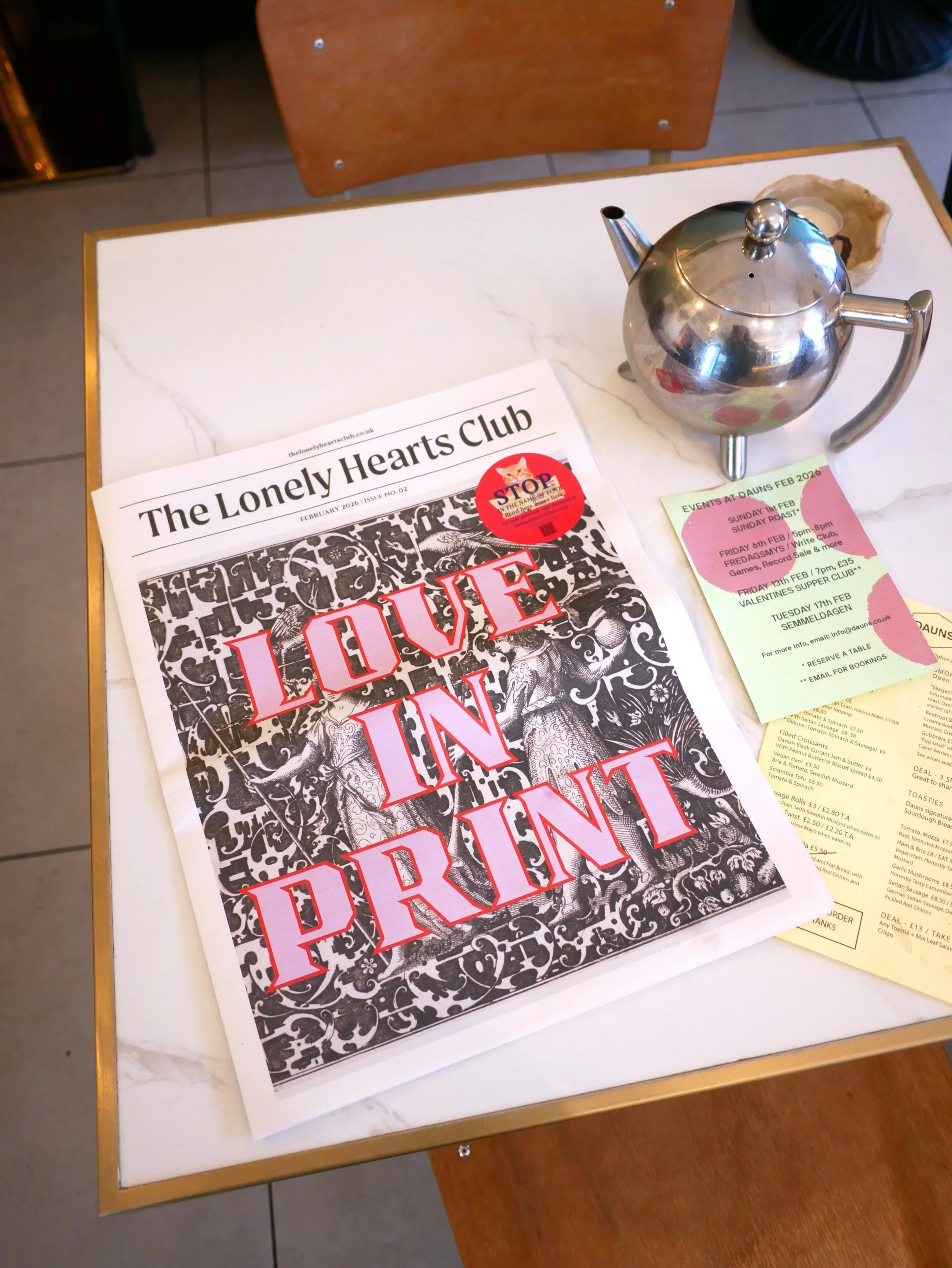 The Lonely Hearts Club Newspaper Issue 02 on a white table with a silver teapot at Dauns Cafe in East London.
