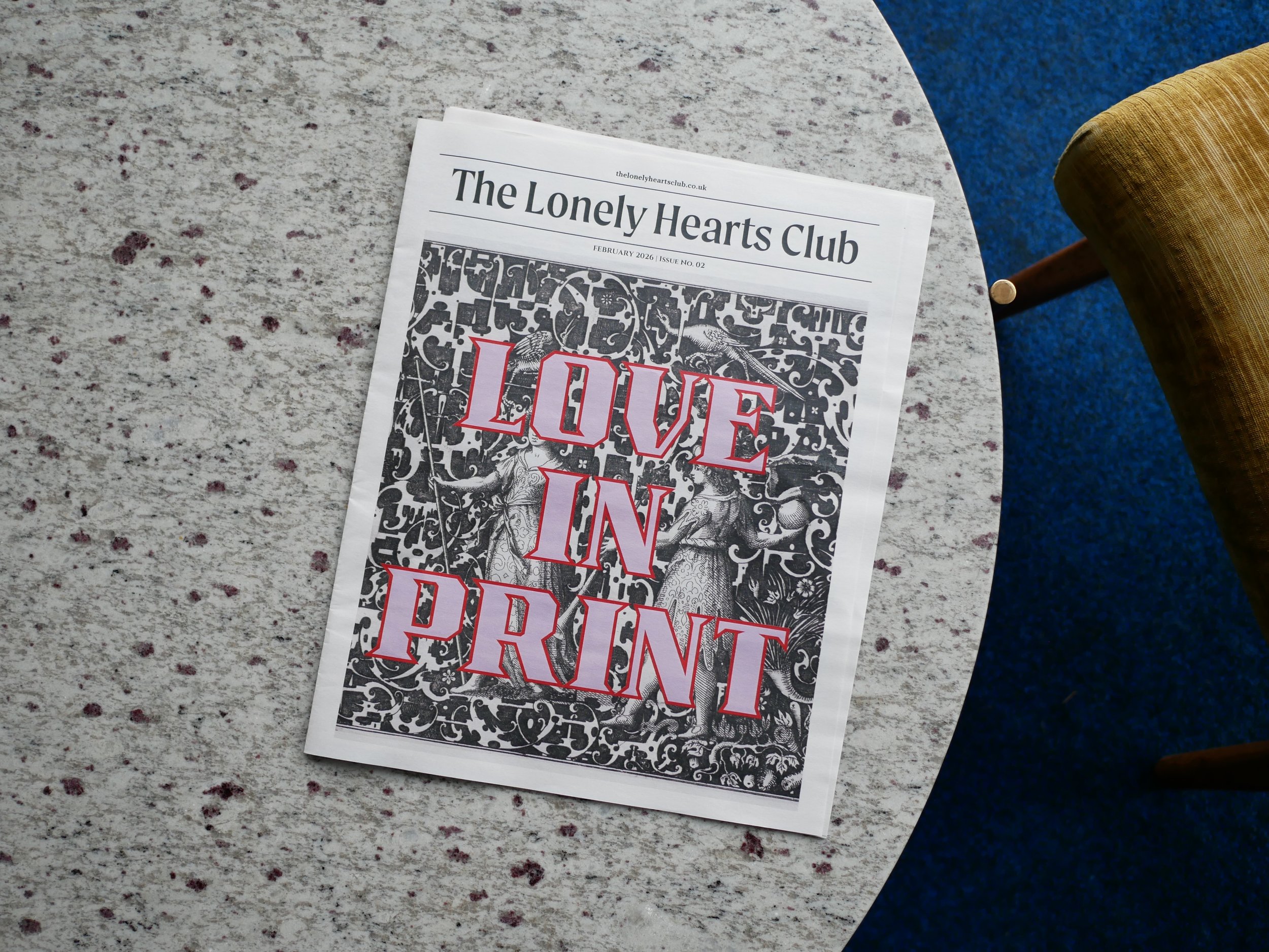 The Lonely Hearts Club Newspaper Issue 02 Cover Page on top of a Marble Table