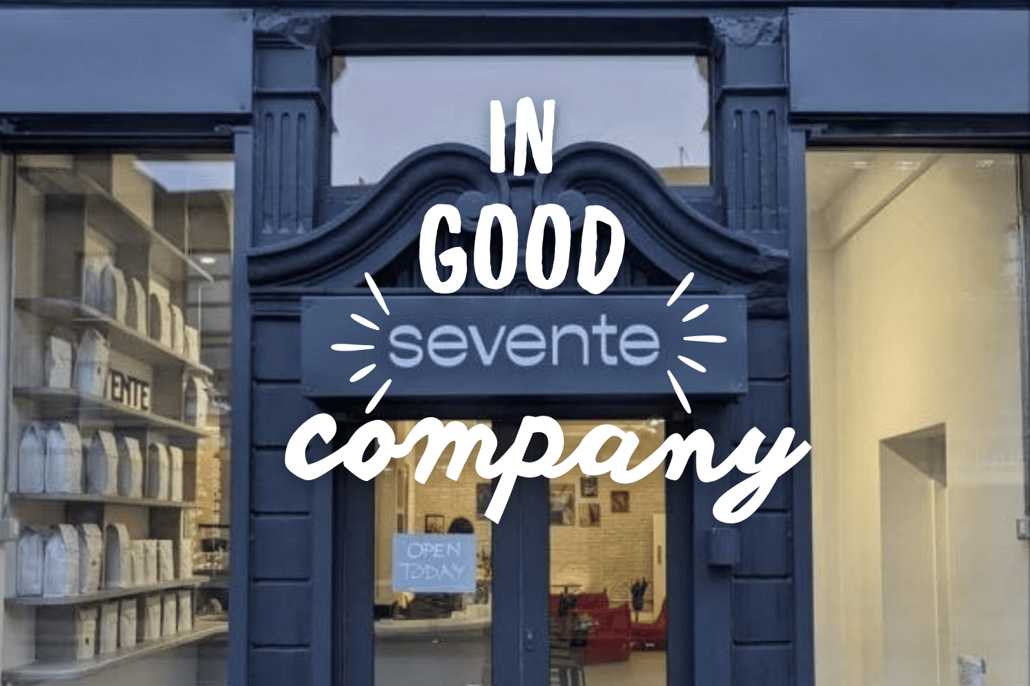 The main door of "Devente" with coffee in the left corner as seen through the window and a little "open today" sign in the door. Title of "In Good Company"  in white script letters.