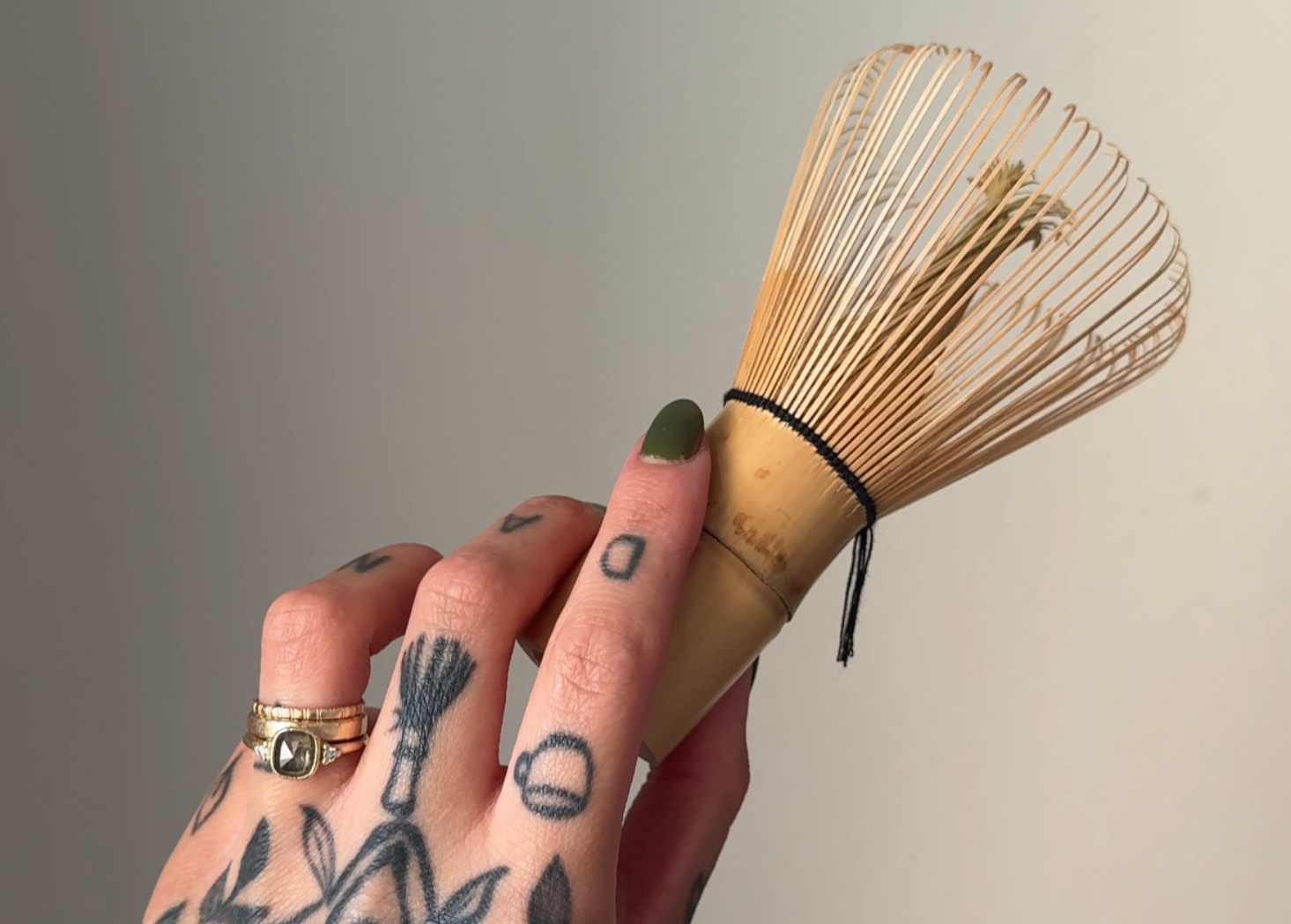 Tattooed hand holding a bamboo whisk (chasen) against a white wall