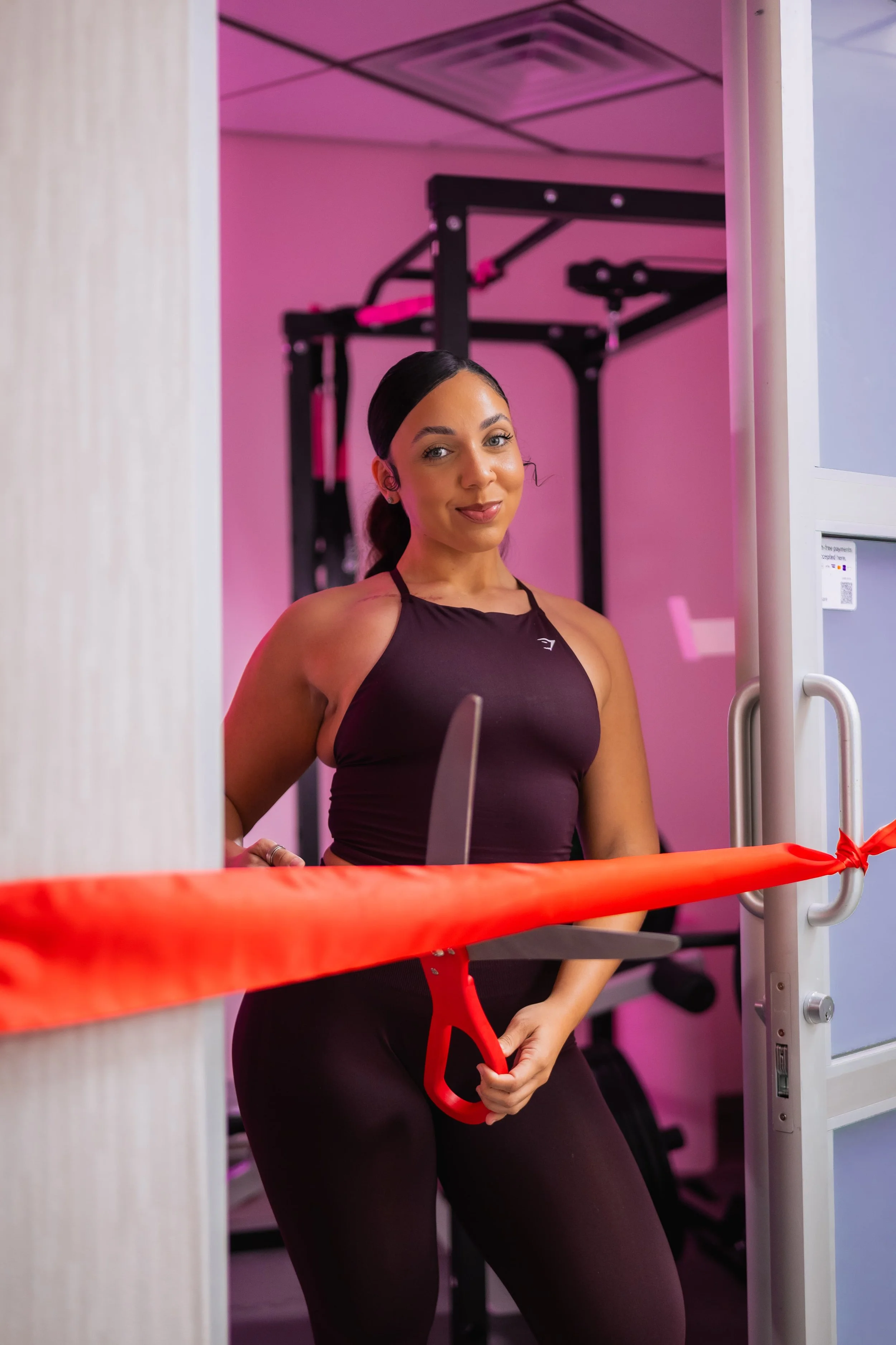 A woman in workout attire holding scissors near a red ribbon in a gym or fitness facility.
