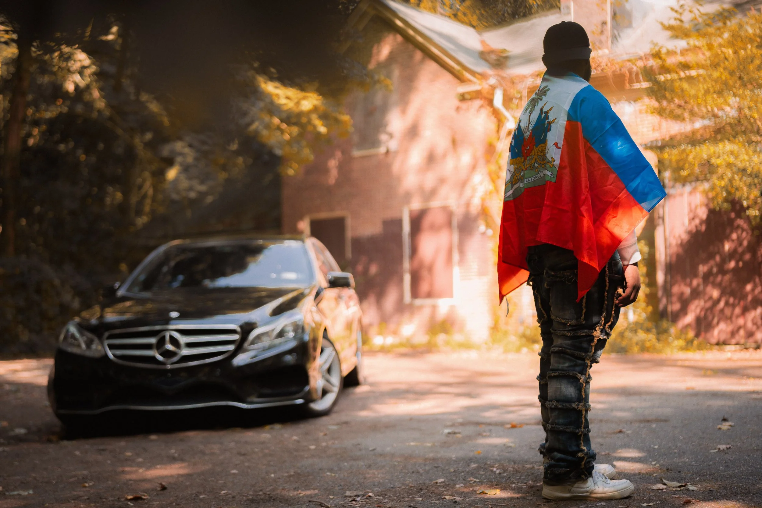 A person standing on the street with an Haiti flag draped over their shoulders, facing a black Mercedes-Benz car, with a house and trees with autumn foliage in the background.