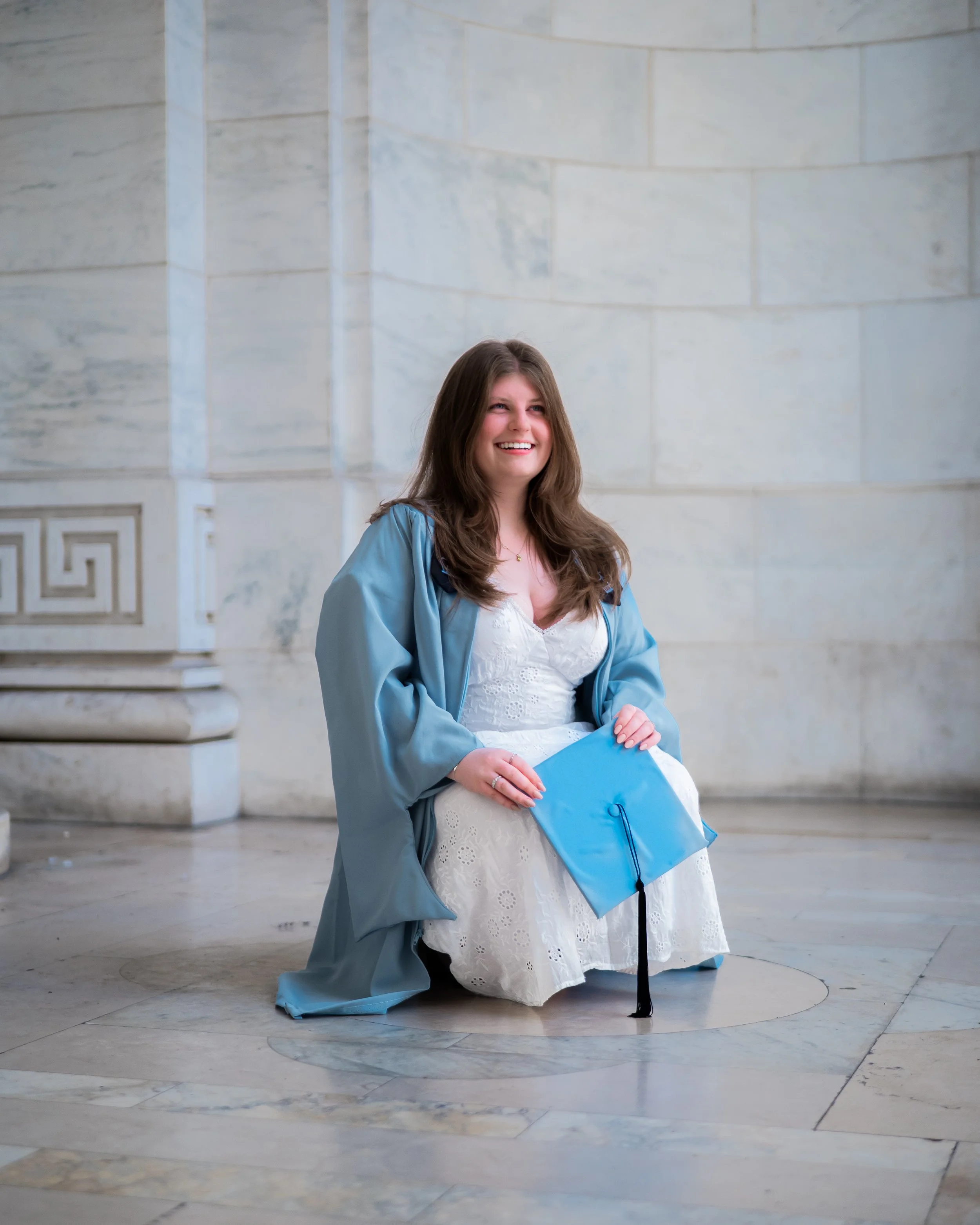 A young woman in a white dress and blue graduation gown kneels on the floor holding a blue graduation cap with a black tassel, smiling indoors against a marble wall