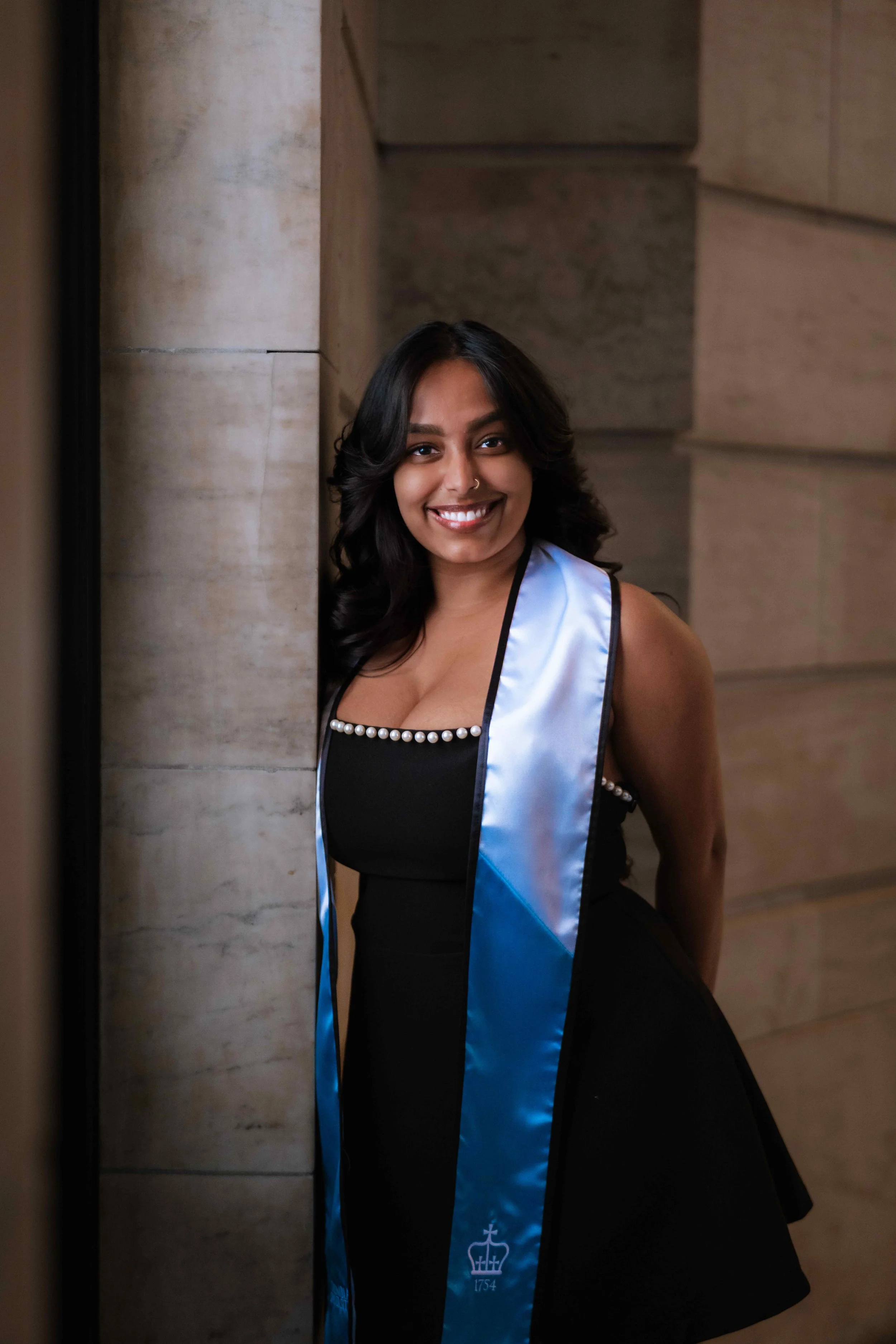 A young woman with black hair, smiling, wearing a black dress with pearl embellishments and a blue satin graduation stole, standing against a stone wall.