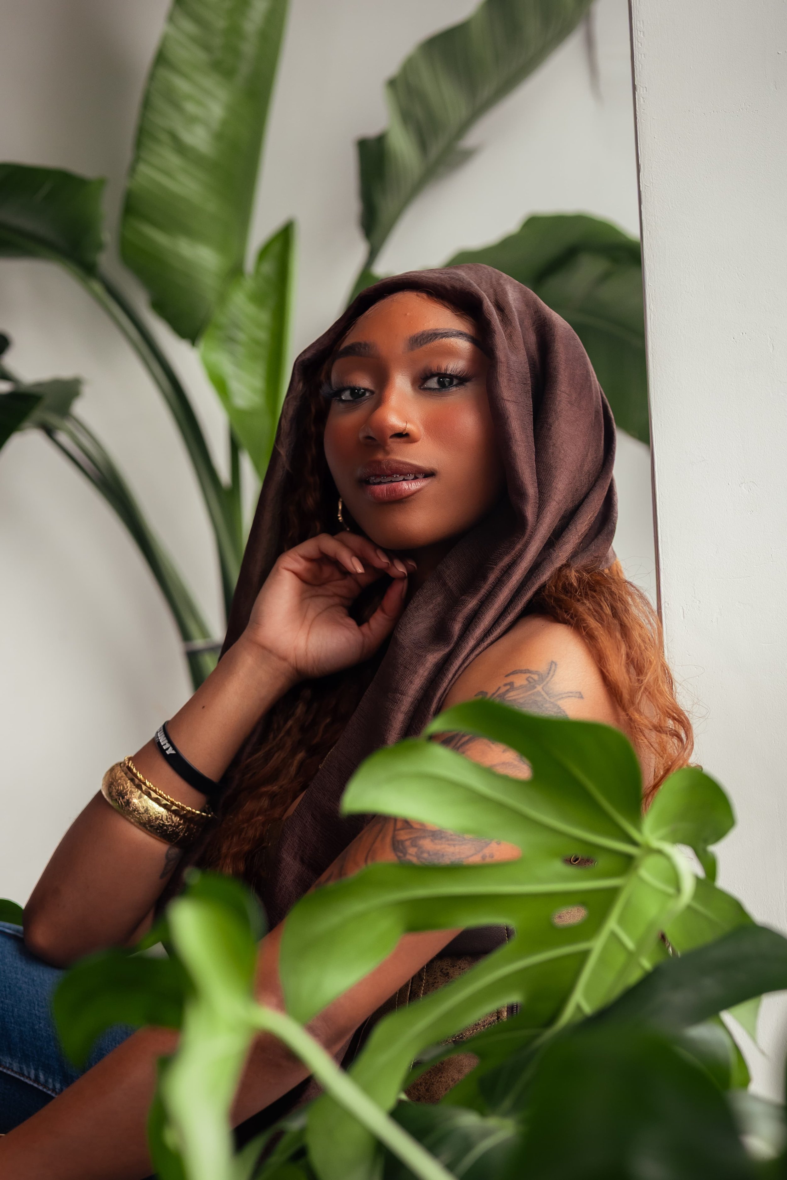 A young woman with long curly hair wearing a brown hooded scarf, gold bracelets, and a tattoo on her upper arm, posing near green tropical plants indoors.