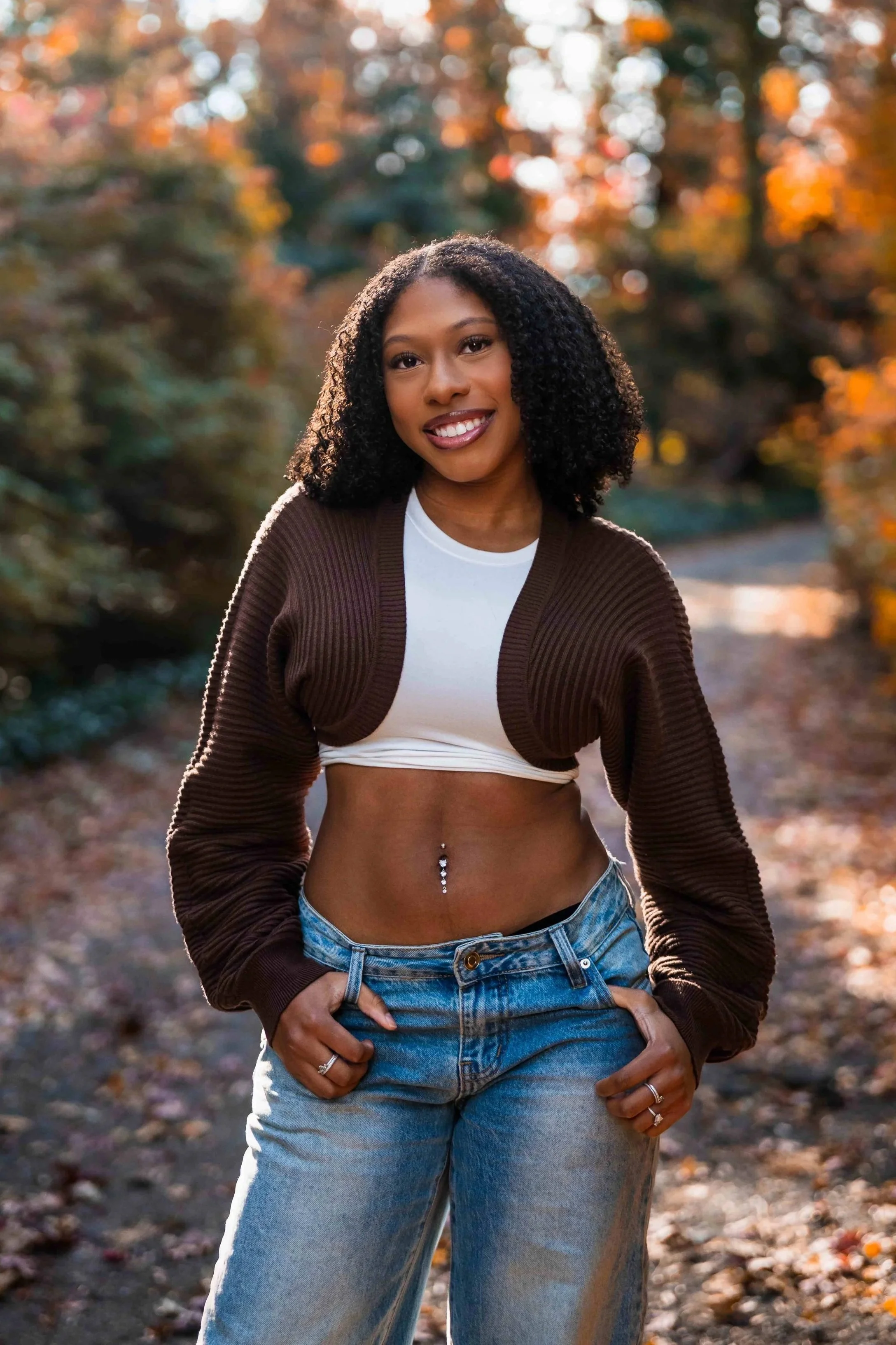 A woman with curly black hair, wearing a white crop top, a brown cardigan, and blue jeans, standing outdoors in a fall setting with trees and orange leaves in the background, smiling and looking at the camera.