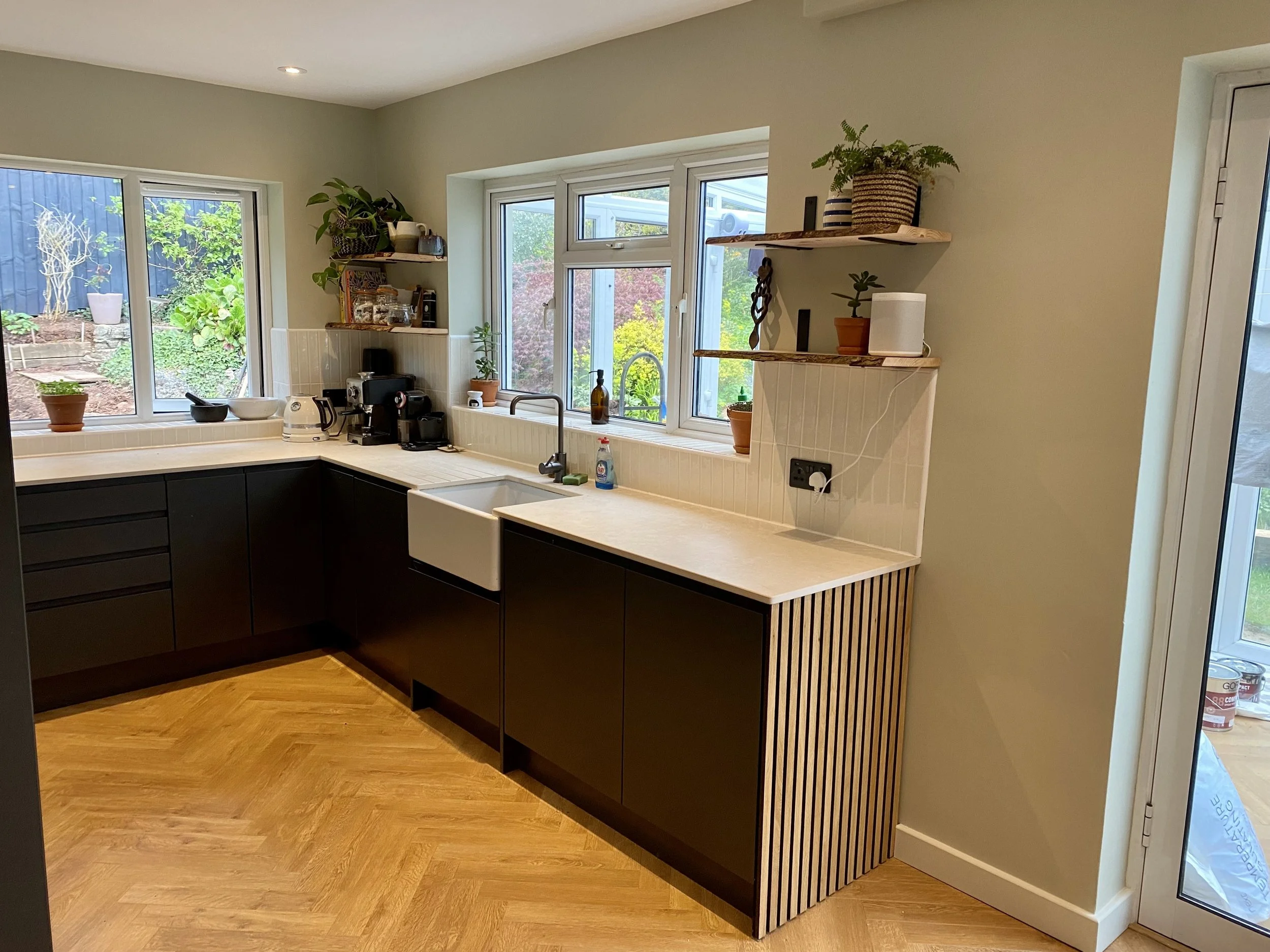 Modern kitchen with black cabinets, white countertops, and large windows showing a garden outdoors. Contains small potted plants, shelves, and kitchen appliances.