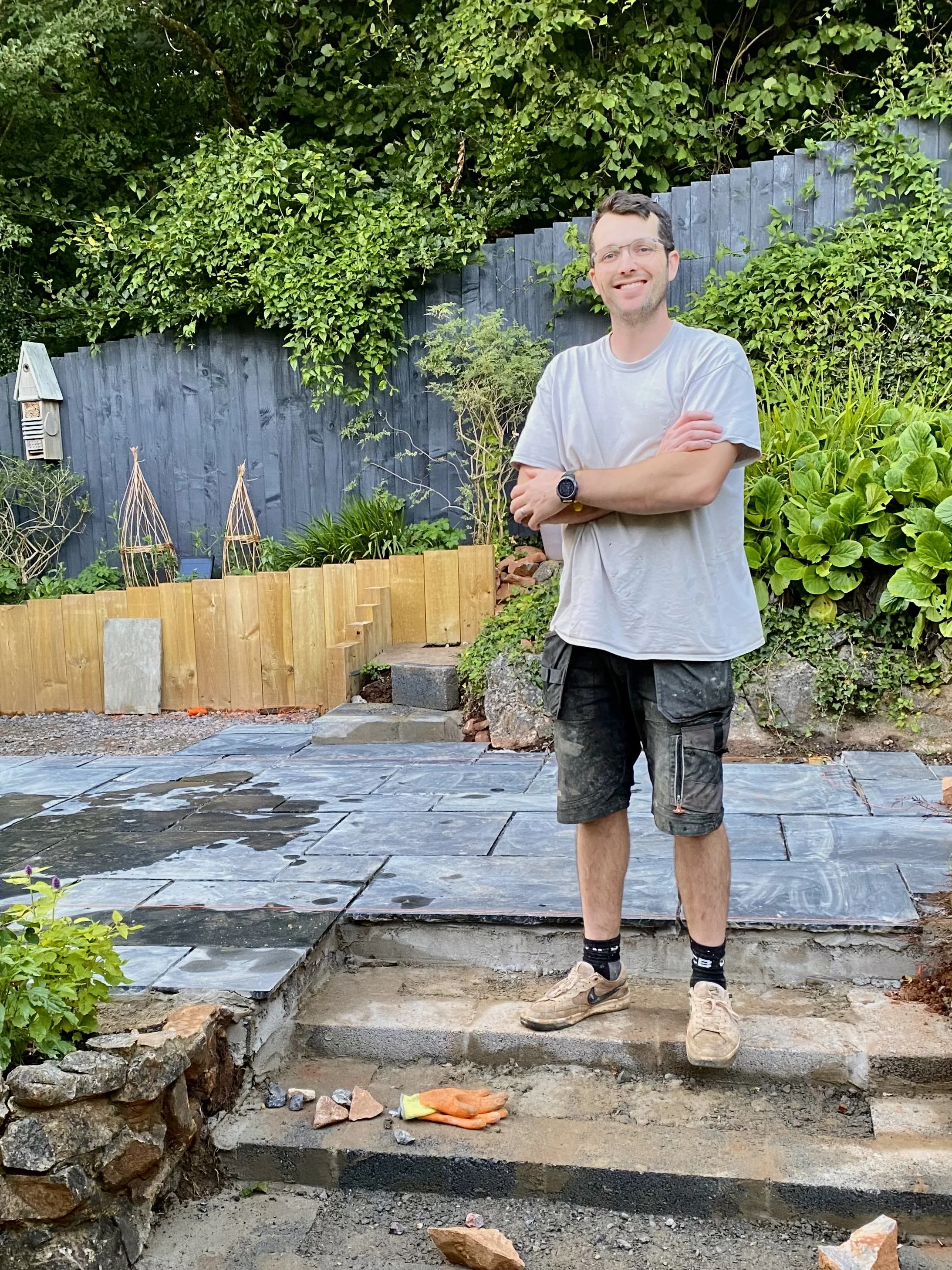 A man standing outdoors in a garden, smiling with arms crossed, wearing a light gray t-shirt, cargo shorts, sneakers, and a watch, with an area under construction or renovation with building materials and tools visible in the foreground.