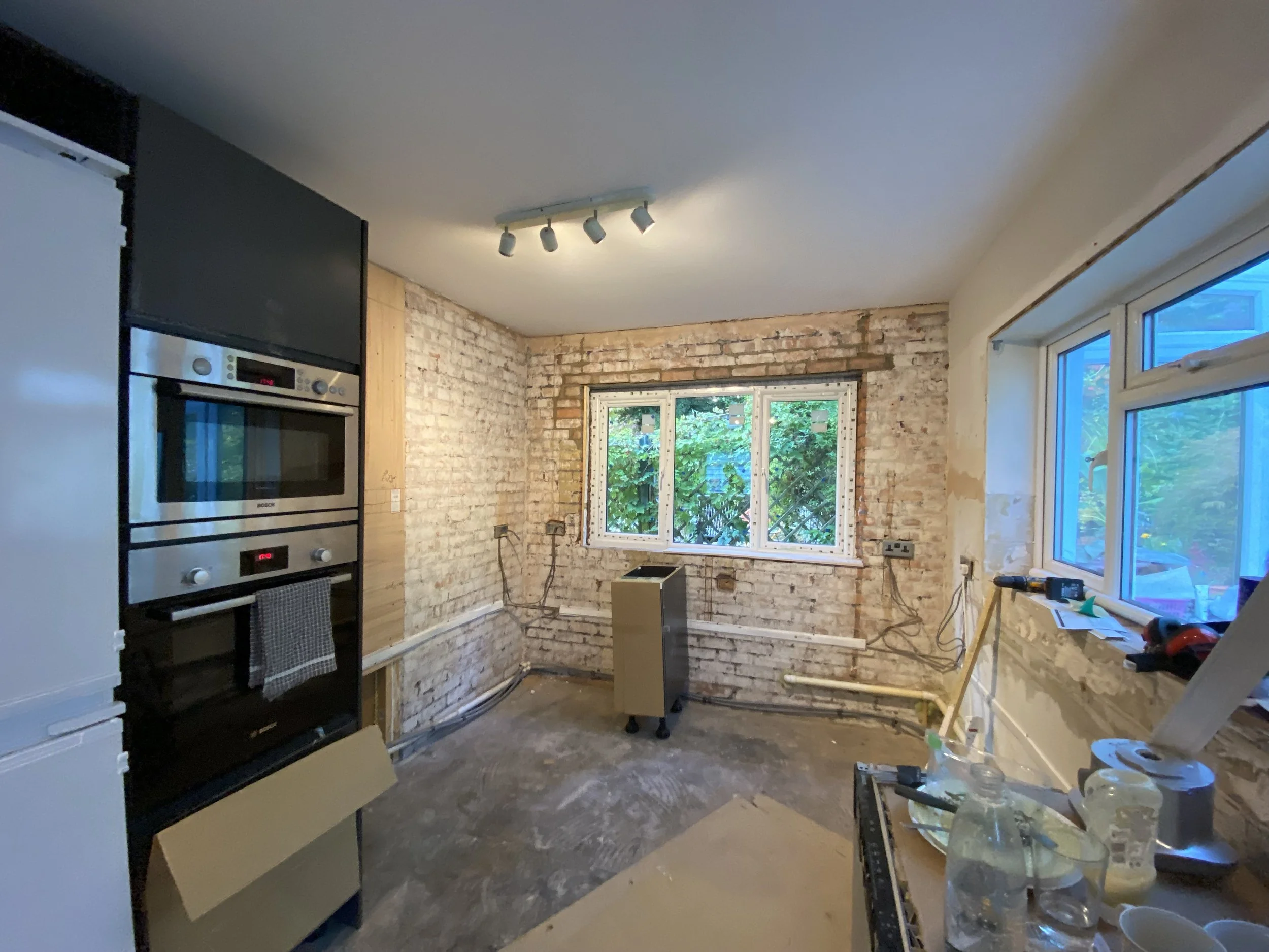 Kitchen undergoing renovation with exposed brick walls, partially installed appliances, and construction tools and materials scattered around.