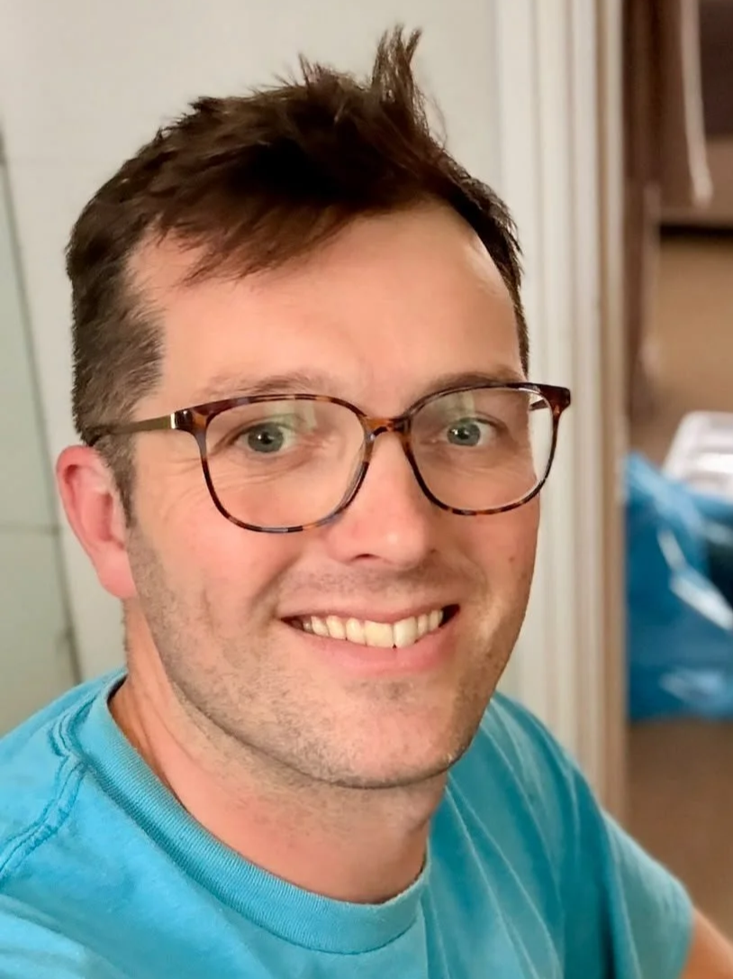 A smiling man with glasses and short brown hair, wearing a blue shirt, looking at the camera in an indoor setting.