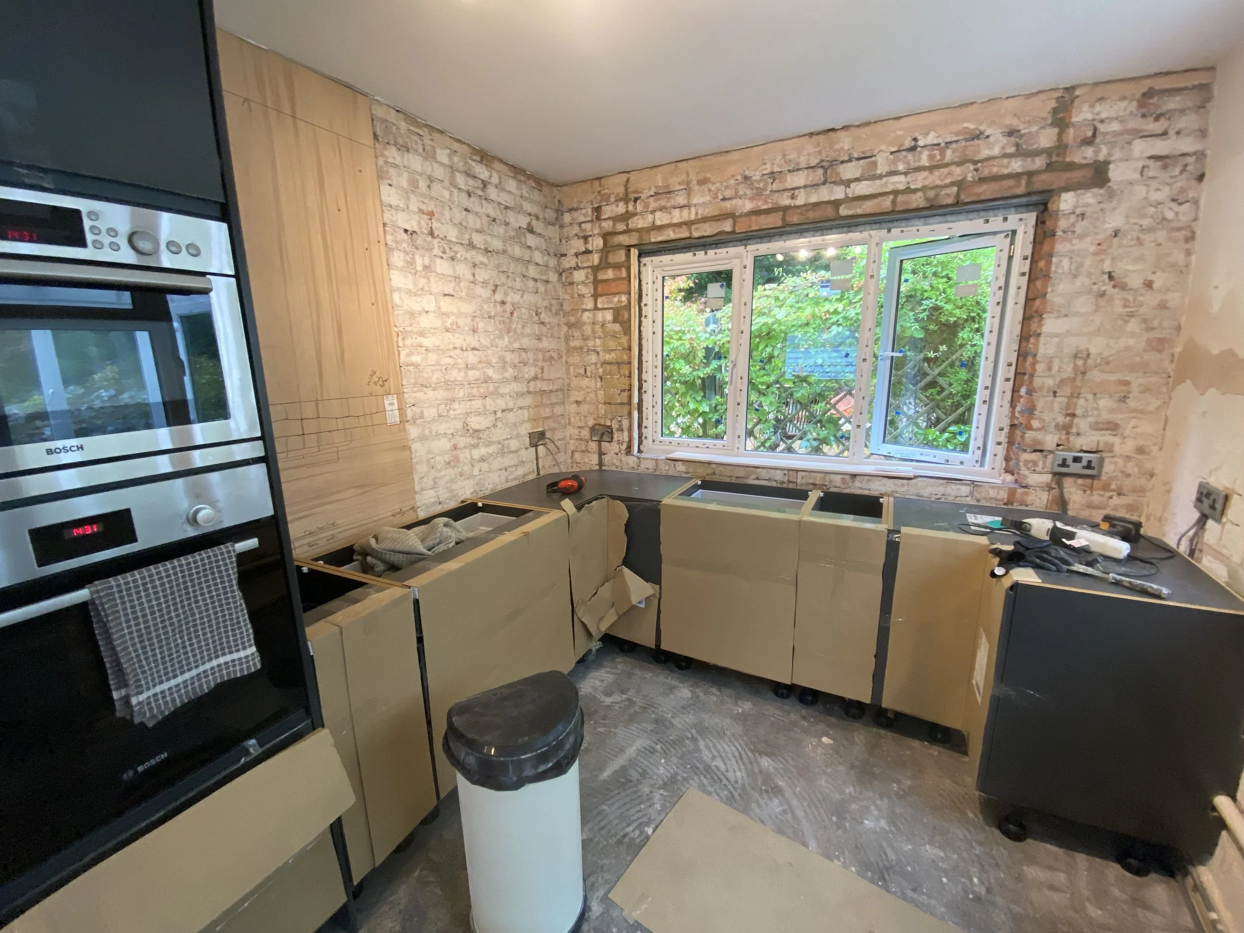 Kitchen under renovation with exposed brick walls, partially installed black cabinets, an oven, and a window overlooking greenery.