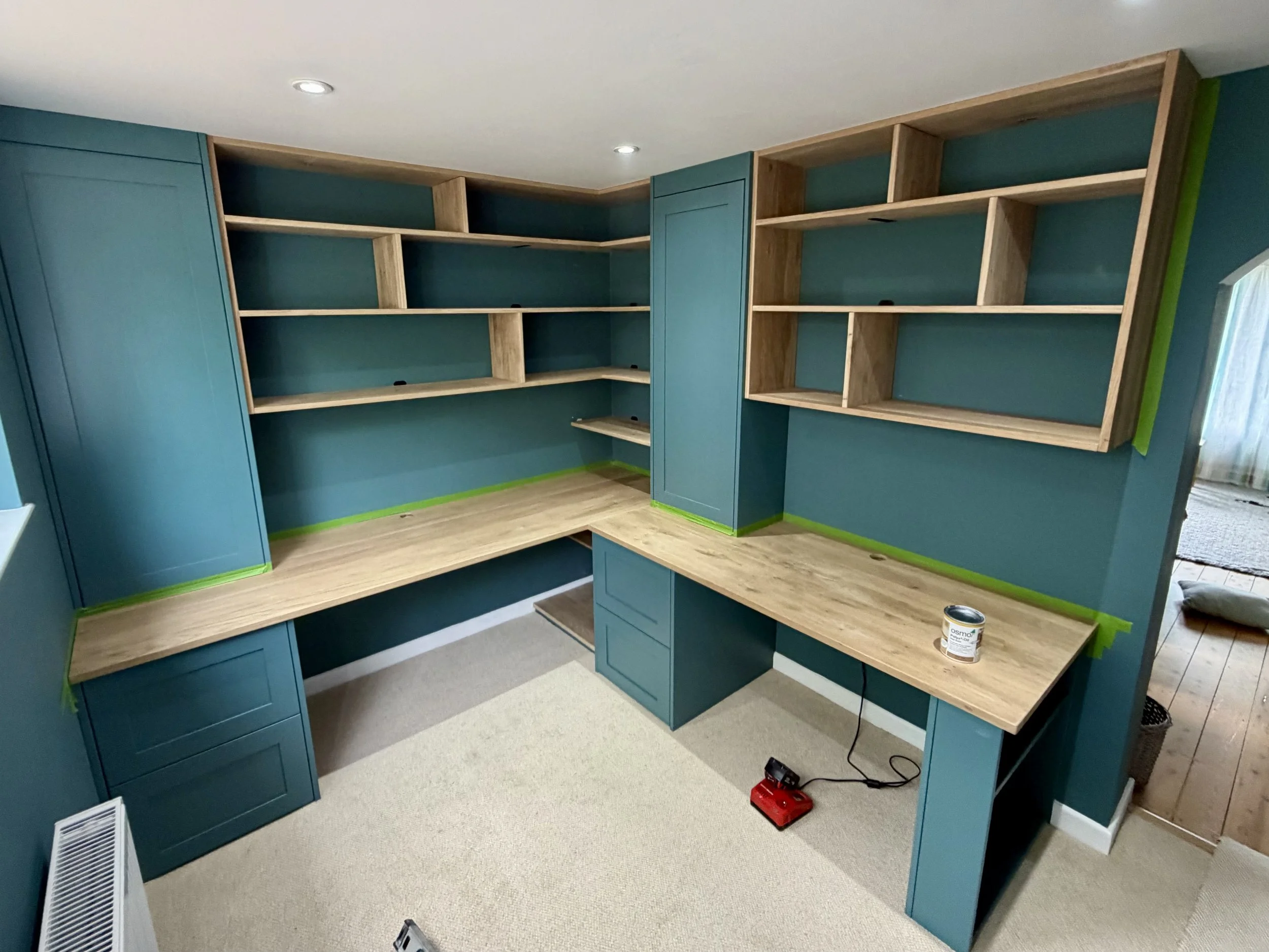 A room with teal built-in shelves and cabinets, some shelves are unfinished with visible wood, a desk area with a large surface and a paint can, a light-colored carpet, and a small window with curtains.