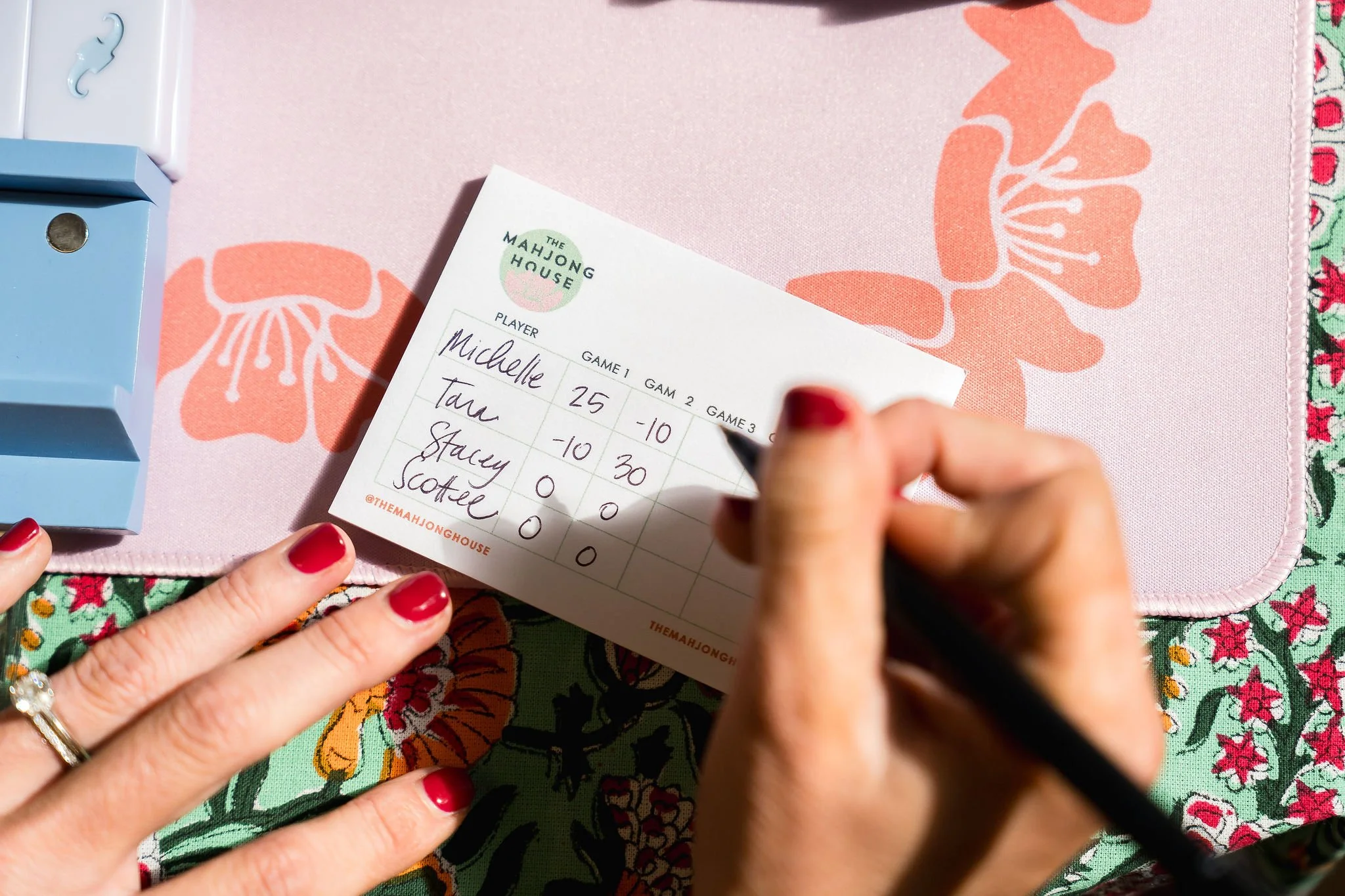 A person with red-painted nails is writing on a scorecard for The Mahjong House, sitting on a pink floral tablecloth with a light blue box nearby.