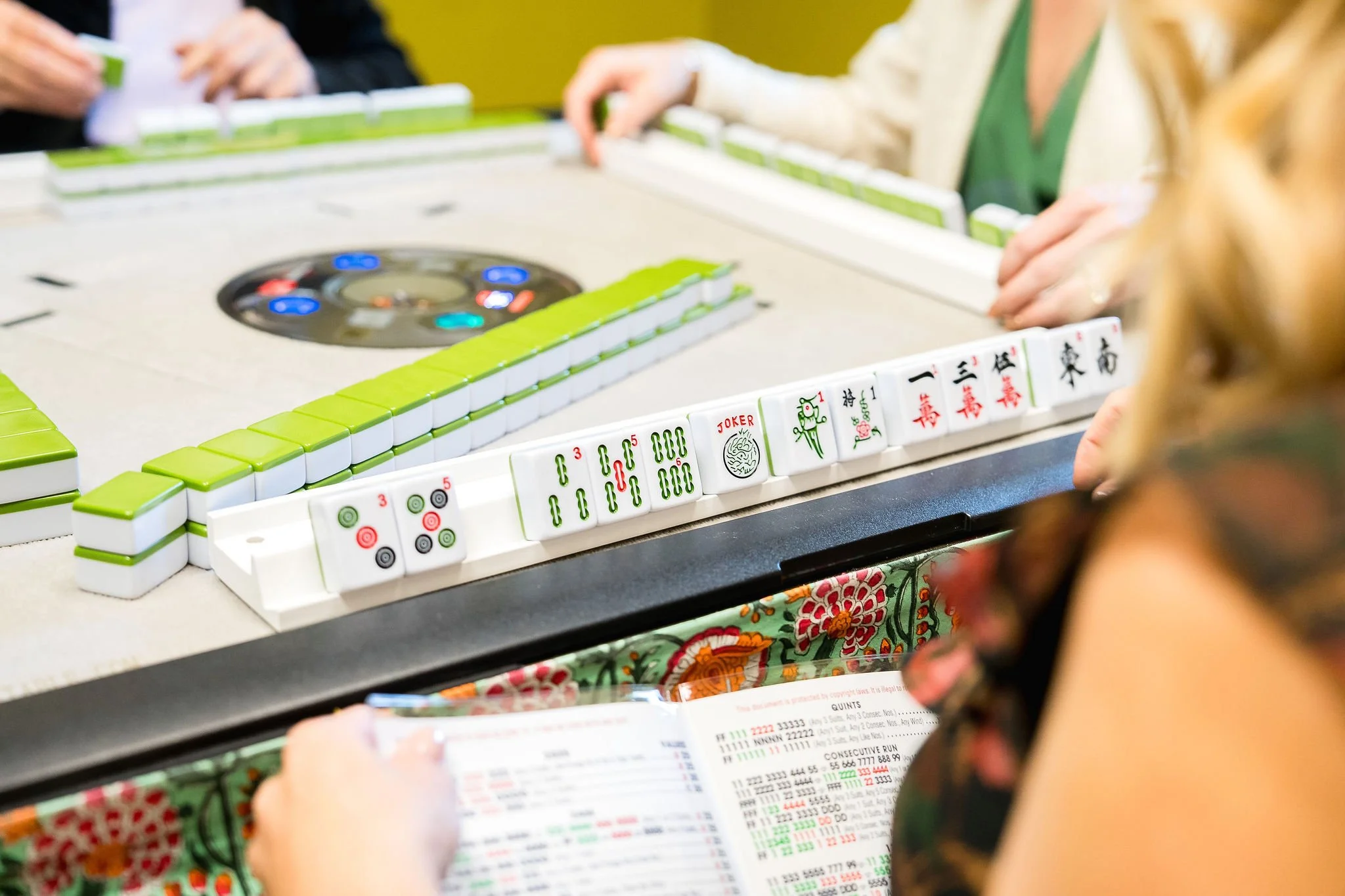Close-up of a mahjong game in progress with players around the table, mahjong tiles and a game control disc visible, and a person reading a score sheet.