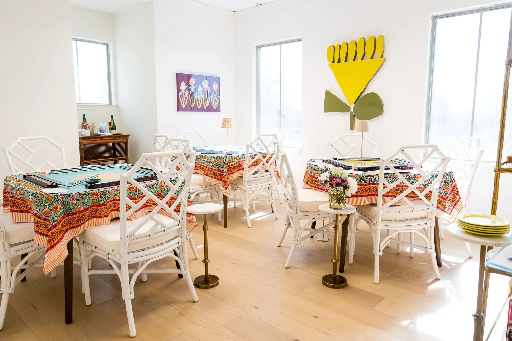 Bright dining room with white chairs and tables covered with colorful, patterned tablecloths. There are two windows letting in natural light, with a small flower arrangement on one table. Decor includes vibrant wall art and a large yellow and green abstract sculpture on the wall.