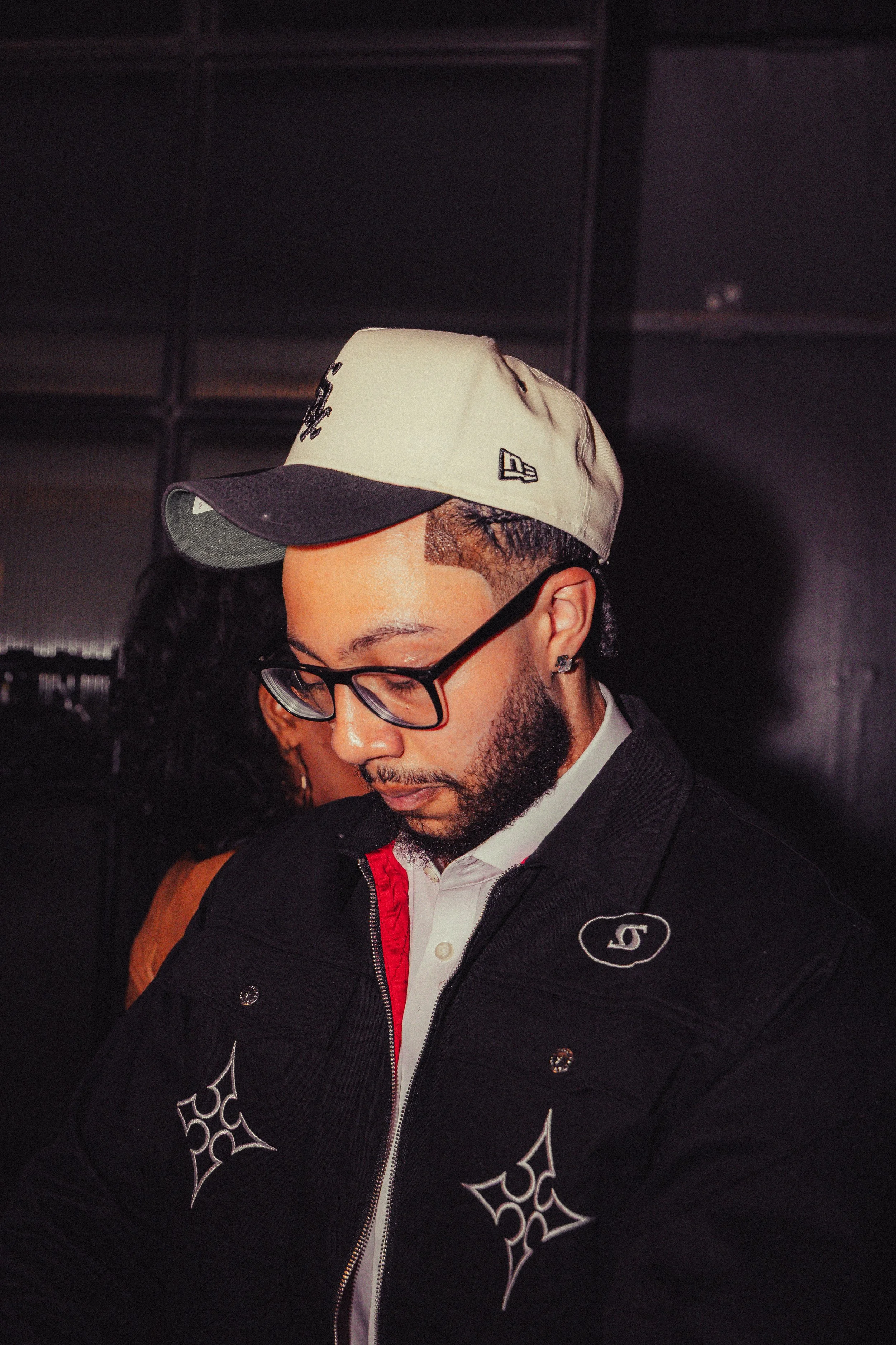 A man with glasses, wearing a beige and black cap, a white collared shirt, and a black jacket with embroidered designs, is looking downward.