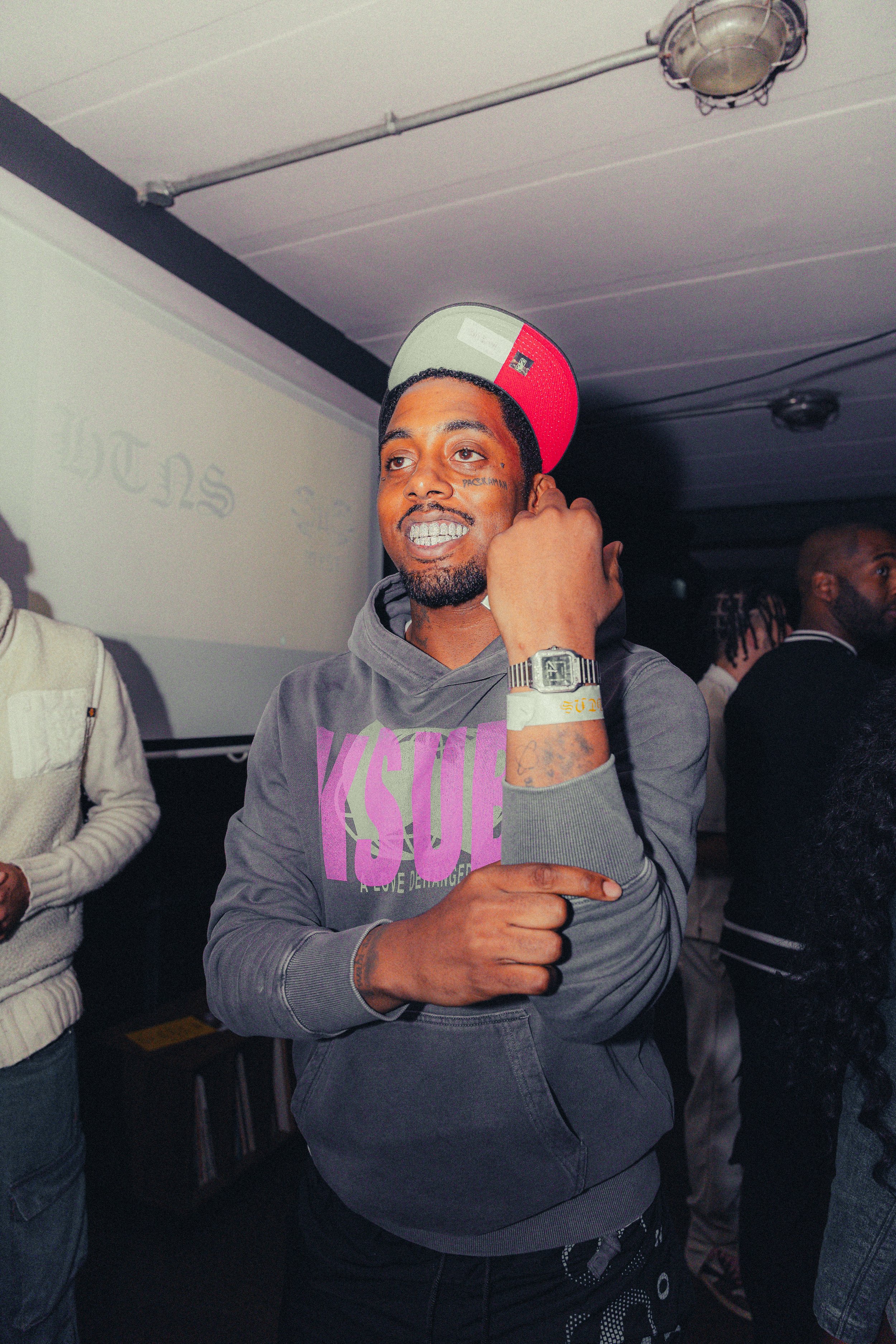 A young man with tattoos, wearing a gray hoodie with purple text, a cap resembling the Italian flag, and a watch, standing in a dimly lit room with people in the background.