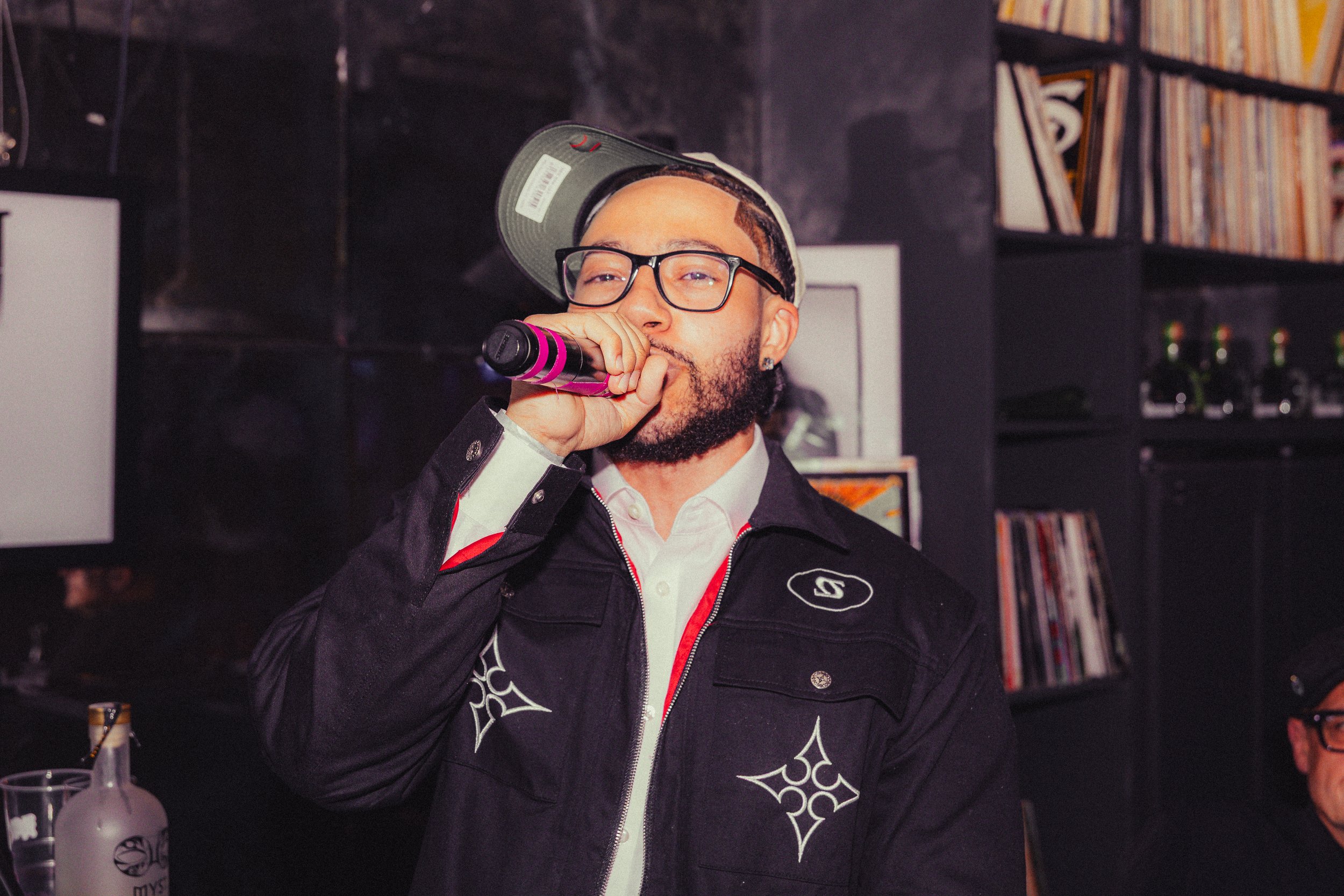 A man with glasses, a beard, and a baseball cap worn backwards is singing into a microphone in a dimly lit room with shelves of records in the background.