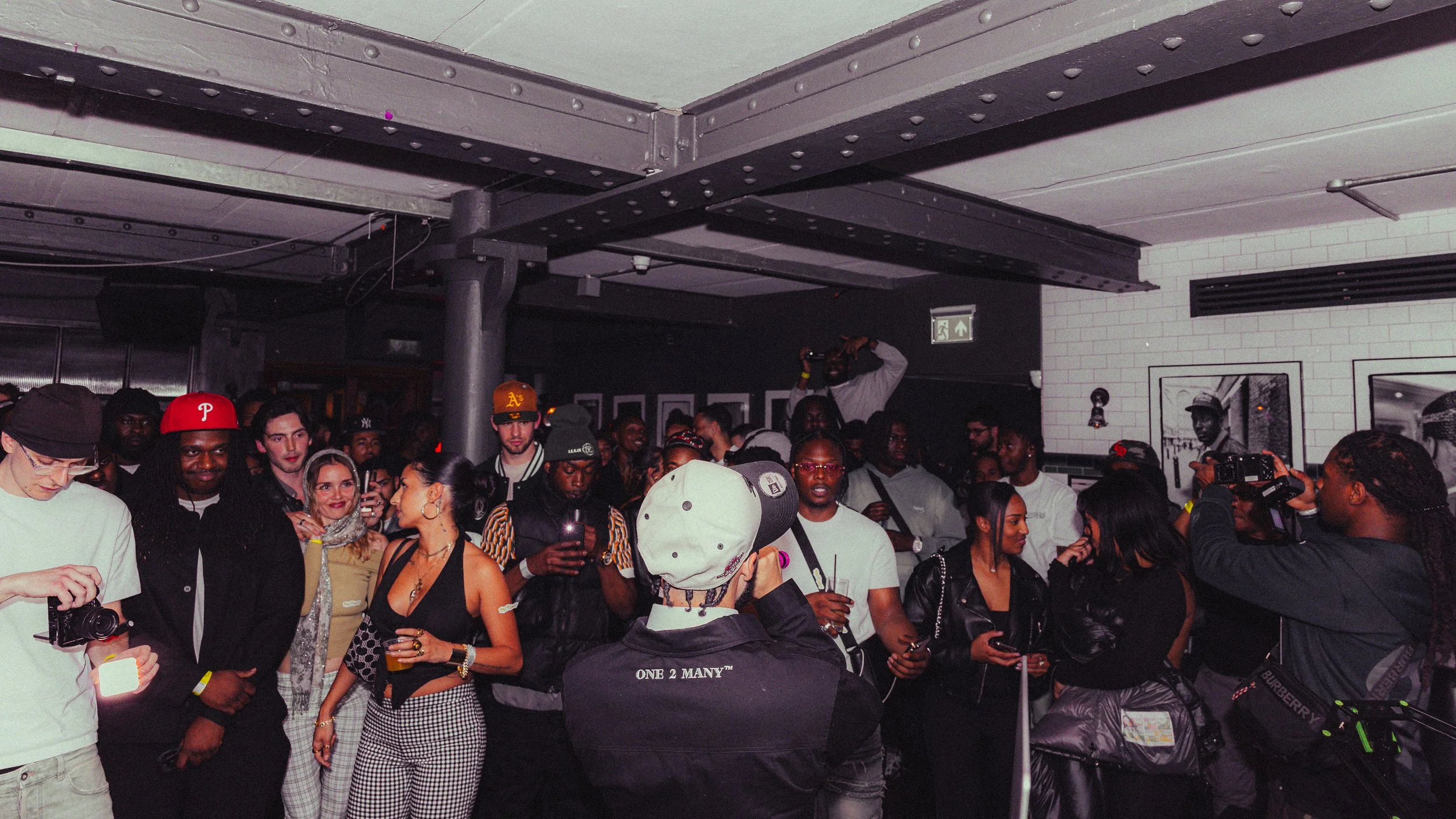 Crowd of people at an indoor social event or party, some taking photos or videos, with a bar or gallery-style setting and black and white photographs on the wall.