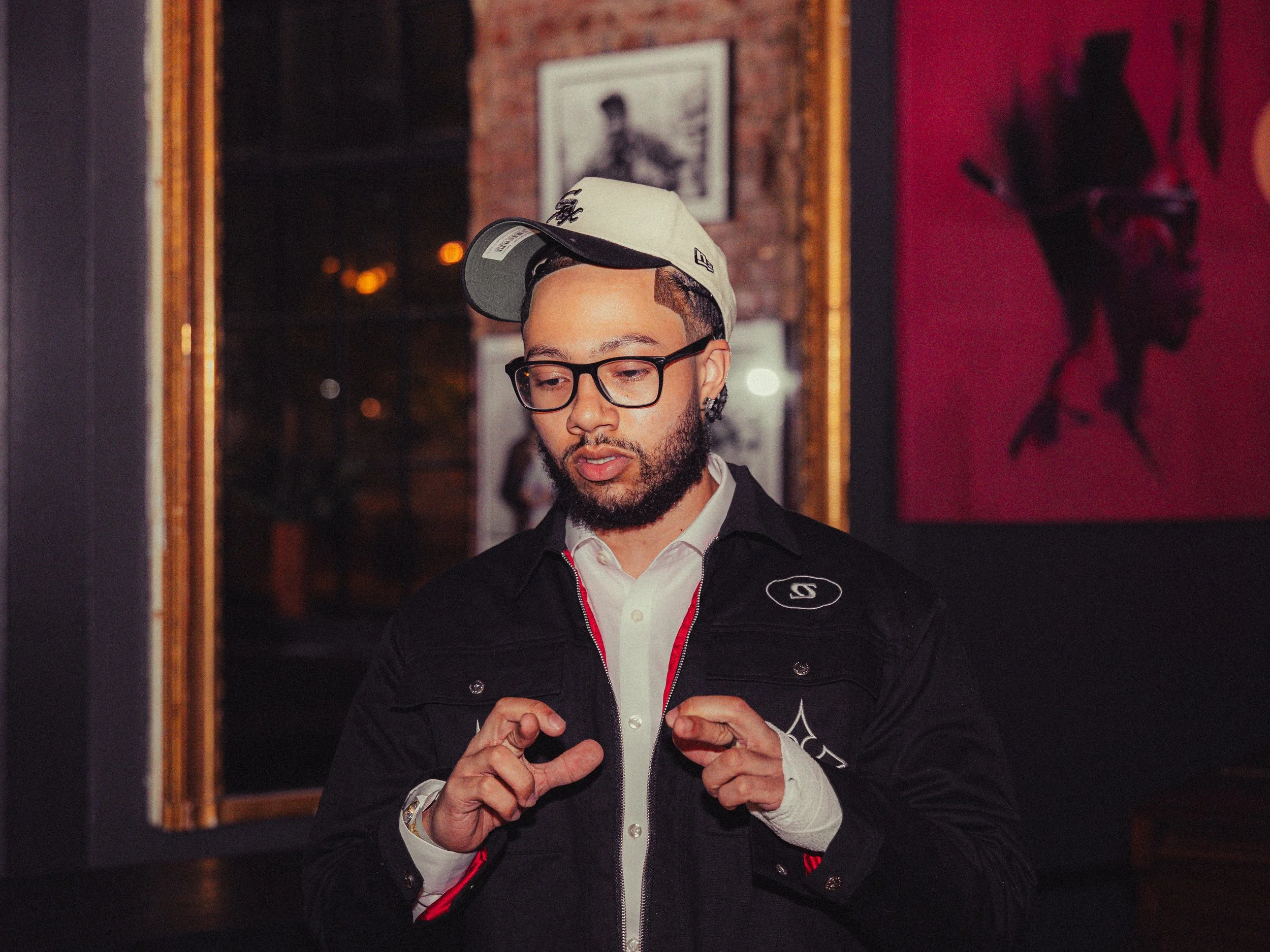 Man wearing glasses, a white polo shirt, a black jacket with logos, and a baseball cap, standing indoors with framed artwork on the wall behind him.
