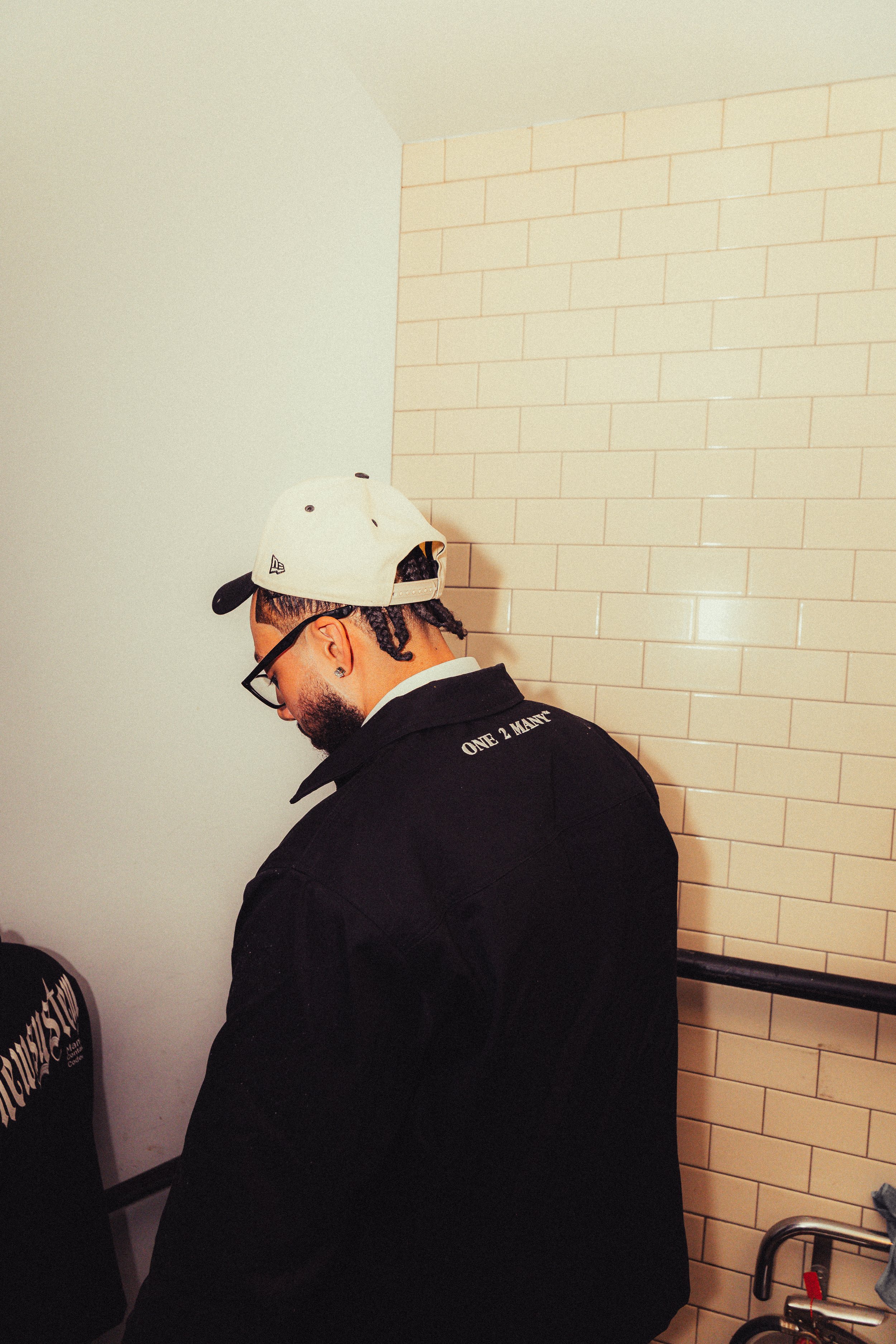 A man with glasses, a beard, and dreadlocks, wearing a beige cap and a black jacket, is standing near a beige tiled wall. The back of the jacket has the words 'ONE 2 MANY' printed on it.