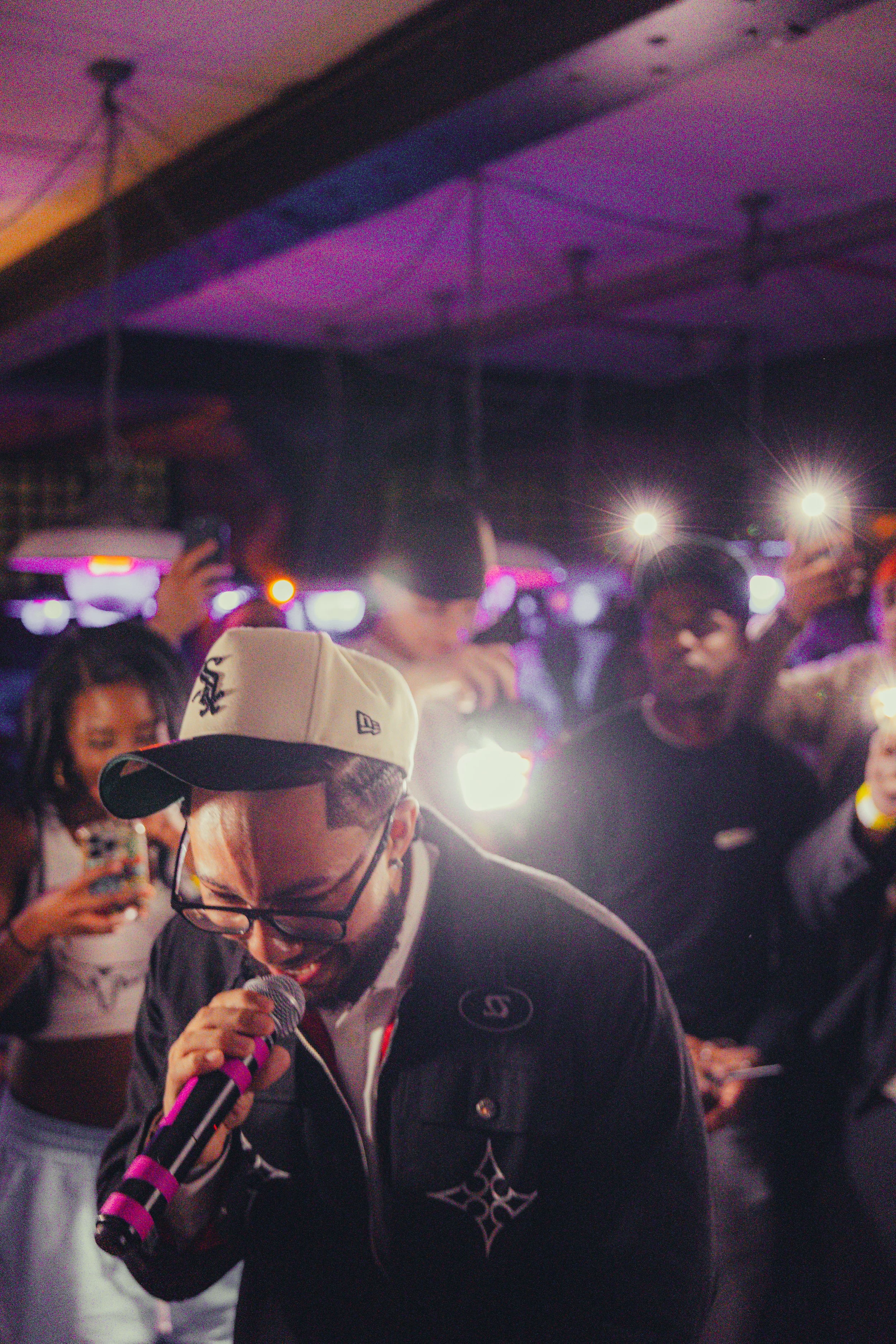 A man with glasses, a beard, and a white cap singing into a microphone at a party or nightclub with people taking photos and bright lights in the background.