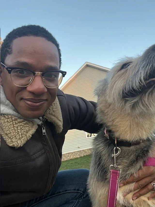 A man wearing glasses and a brown jacket with a fur collar taking a selfie with his large gray dog, outdoors during daytime.