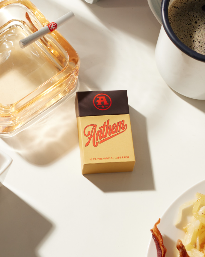 Pack of Anthem pre-rolled cigarettes on a white table with a glass ashtray, a cup of coffee, and a plate with food.
