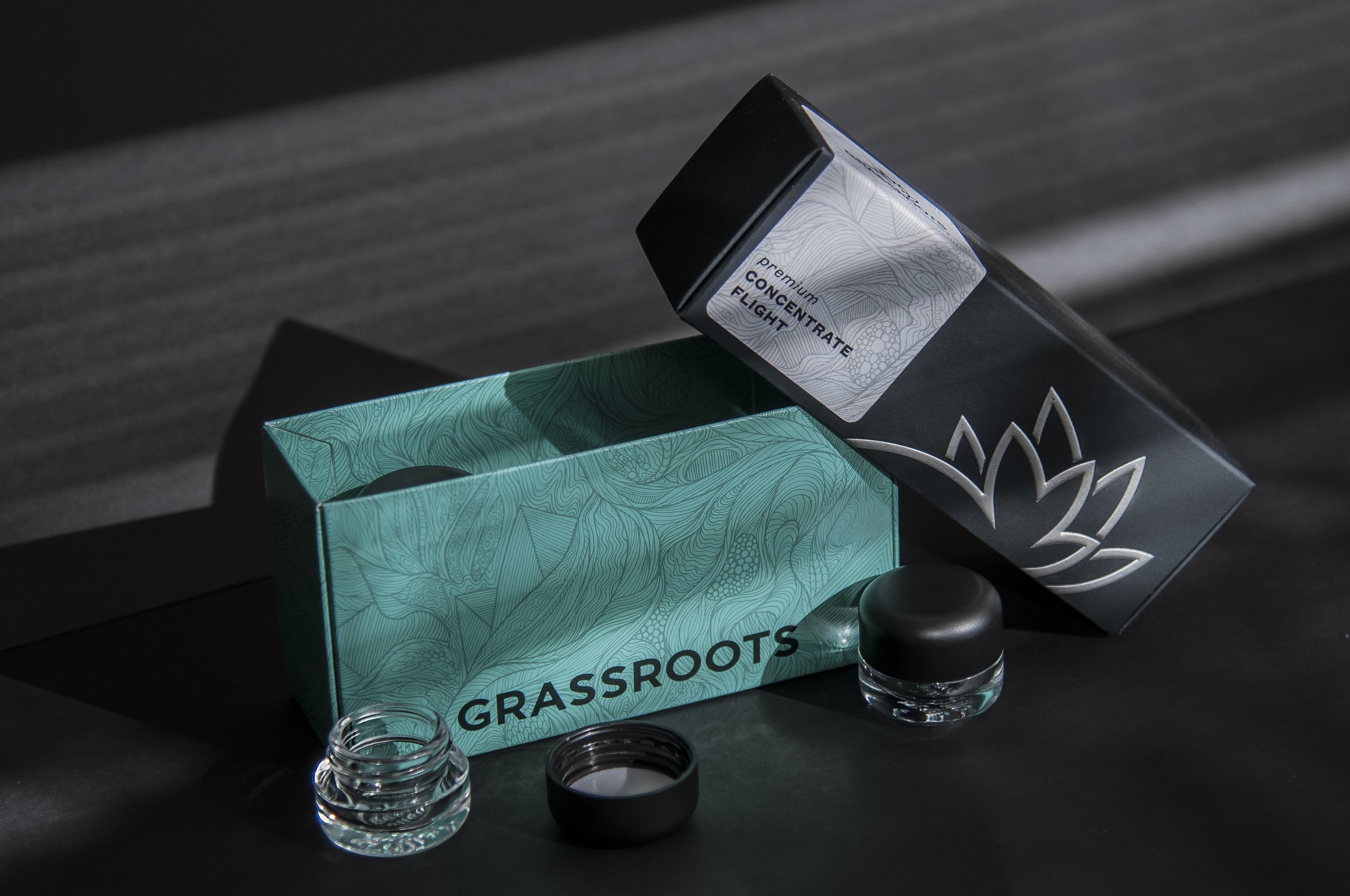 A black box labeled 'Premium Concentrate Flavour' sits on a dark surface with a teal box labeled 'GROUNDS' and a jar of black liquid with a black lid in front of it. Two glass jars are also visible, one in the foreground and one near the teal box.