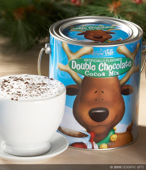 A white cup of frothy hot chocolate topped with cocoa powder, and a tin of Snow Valley Double Chocolate Cocoa Mix with a reindeer on the label, sitting on a wooden surface with blurred green and red holiday decorations in the background.