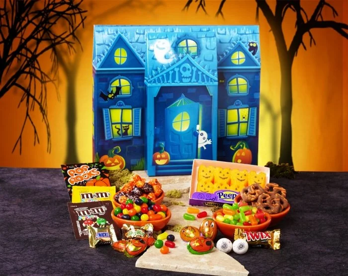 Halloween-themed gingerbread house with ghost, ghost, pumpkin, and bat decorations. Halloween candies, chocolates, and snacks are displayed in front.