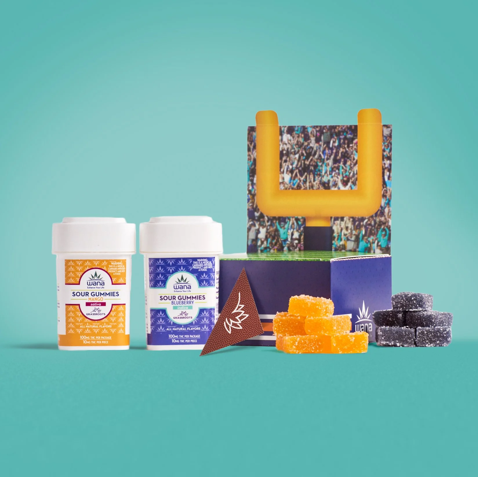 Two containers of WANA sour gummies, one with mango flavor and one with blueberry flavor, a triangle label, a cardboard box with a crowd photo and a yellow cannabis-shaped handle, and assorted gummies on a teal surface.