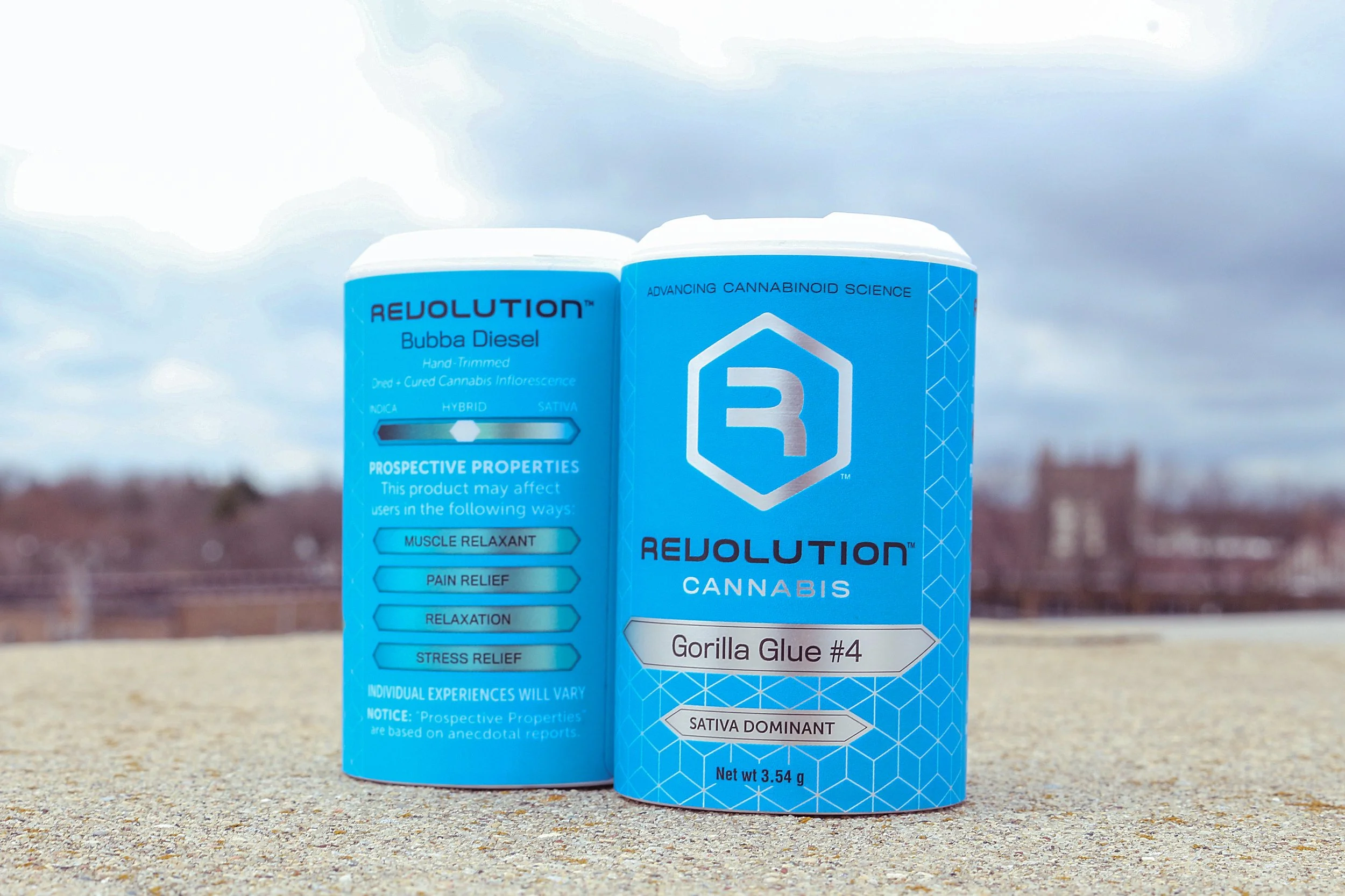 A blue container labeled 'Revolution Cannabis Gorilla Glue #4' with cannabis information and benefits, sitting on a concrete surface outdoors with a cloudy sky and distant buildings in the background.